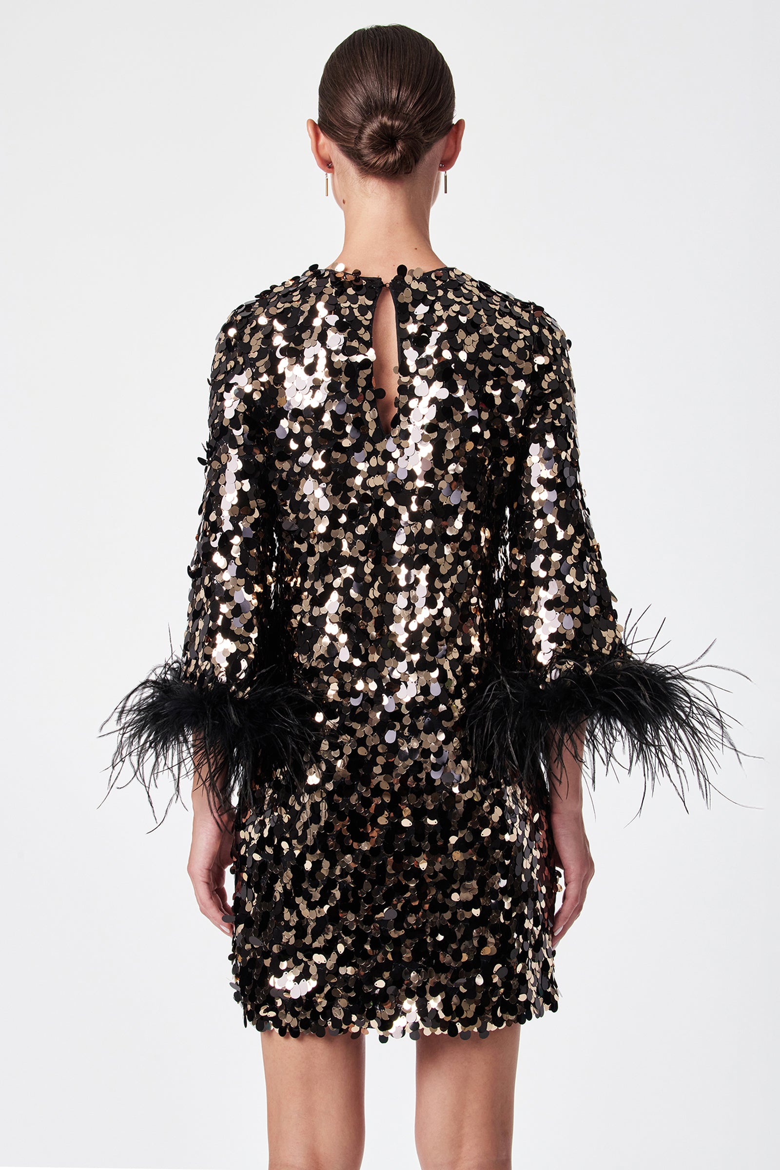 A woman faces away, wearing the Juliet Dress—a short black dress adorned with gold and silver sequins and black feather sleeve trim. Her hair is in a sleek bun, and she stands against a plain white background.