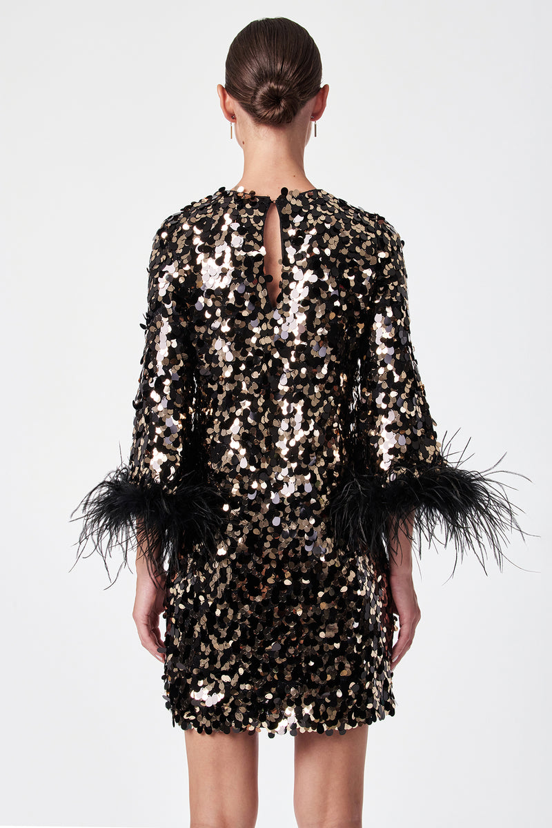 A woman faces away, wearing the Juliet Dressâ€”a short black dress adorned with gold and silver sequins and black feather sleeve trim. Her hair is in a sleek bun, and she stands against a plain white background.