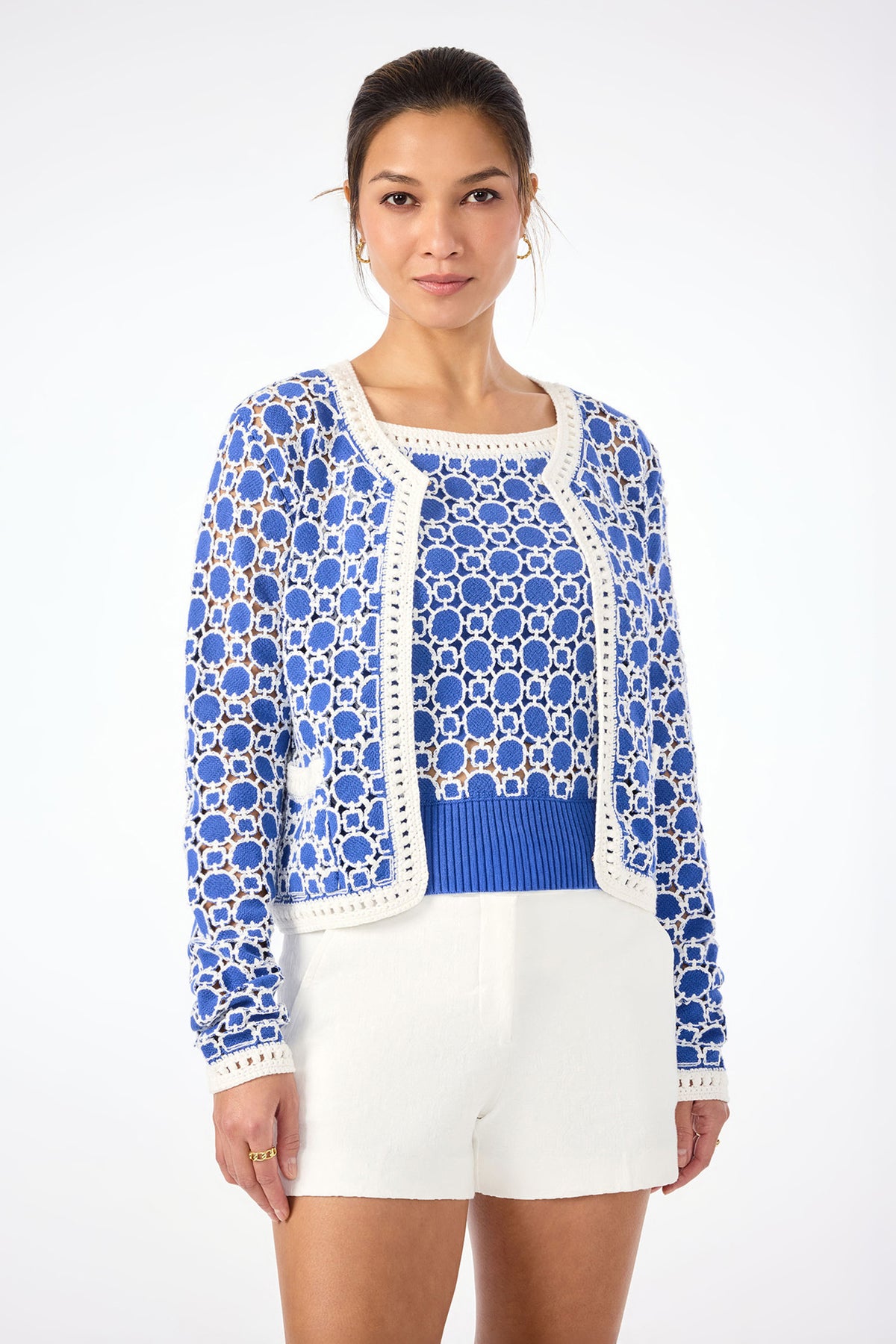 A woman wears the Sinai Contrast Trim Knit Cardigan in Cobalt Cove over a blue top with white shorts, standing against a plain white background. Her hair is tied back, and she gazes confidently at the camera.