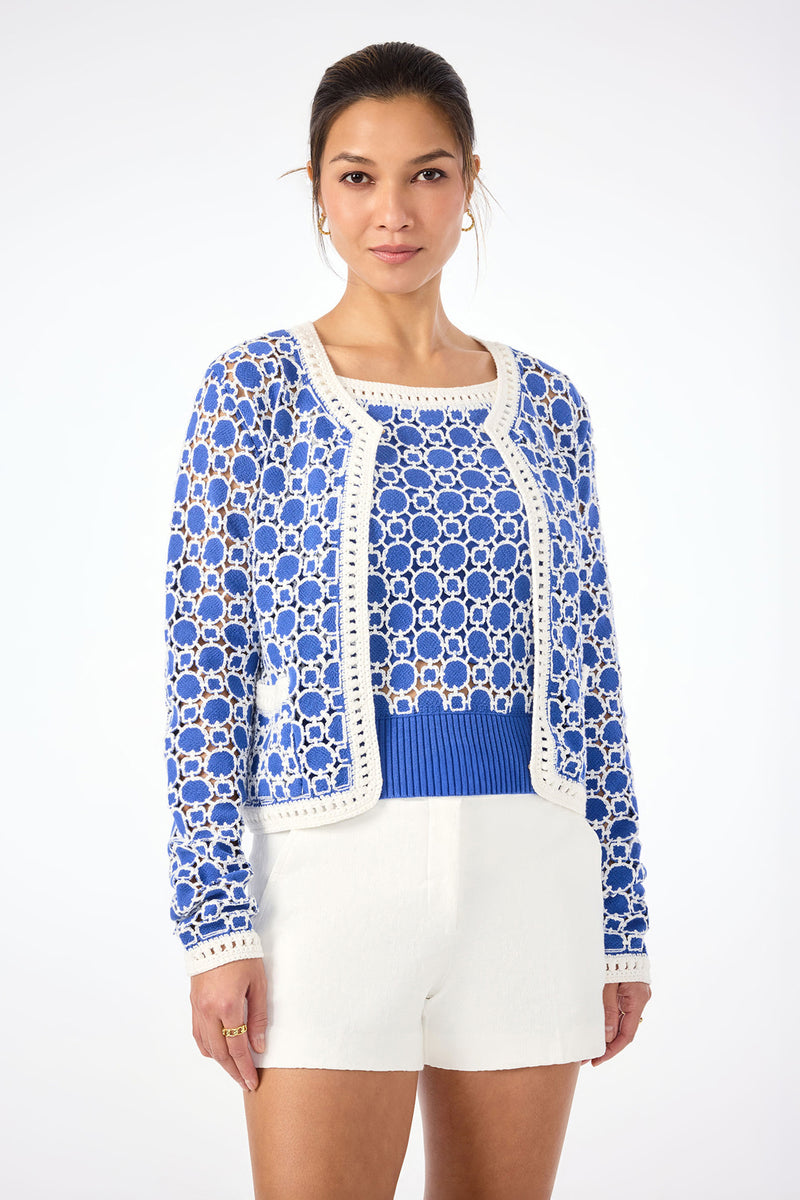 A woman wears the Sinai Contrast Trim Knit Cardigan in Cobalt Cove over a blue top with white shorts, standing against a plain white background. Her hair is tied back, and she gazes confidently at the camera.