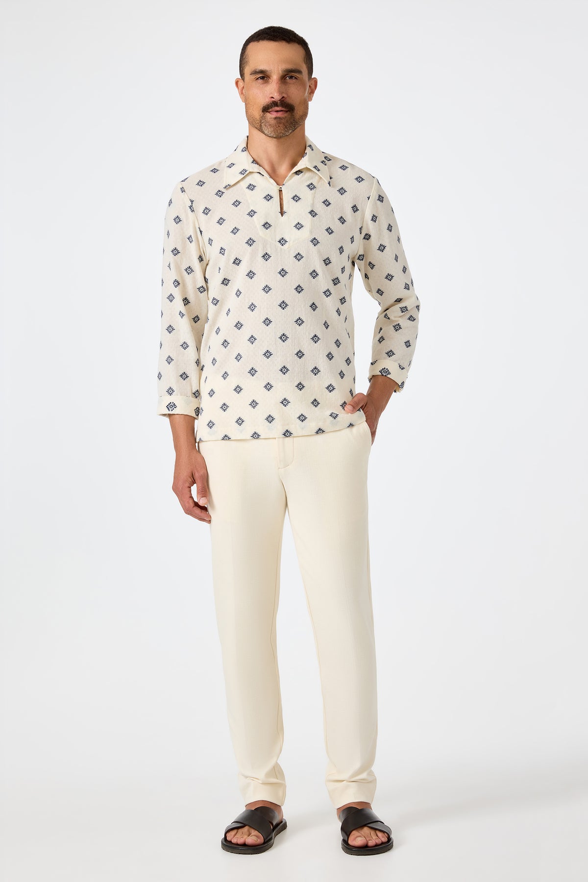 A man with short hair and a mustache stands against a white background, wearing the Hendrix Long-Sleeve Pullover in cream, cream pants, and black sandals. He poses with one hand in his pocket.