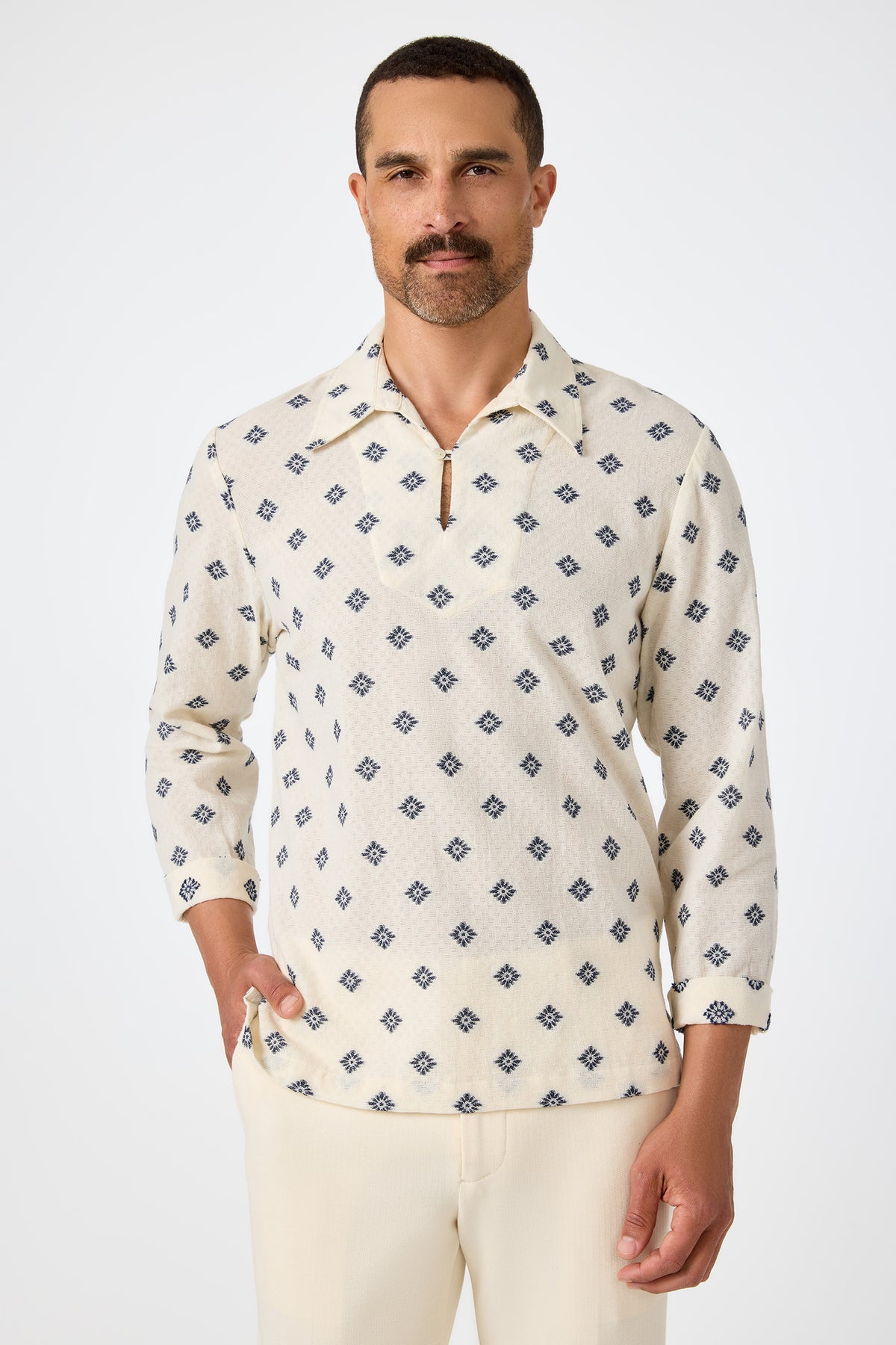 A man with short dark hair and a mustache wears the Hendrix Long-Sleeve Pullover in ecru, featuring a blue geometric pattern and a short zip at the collar, paired with light trousers, standing against a plain white background.