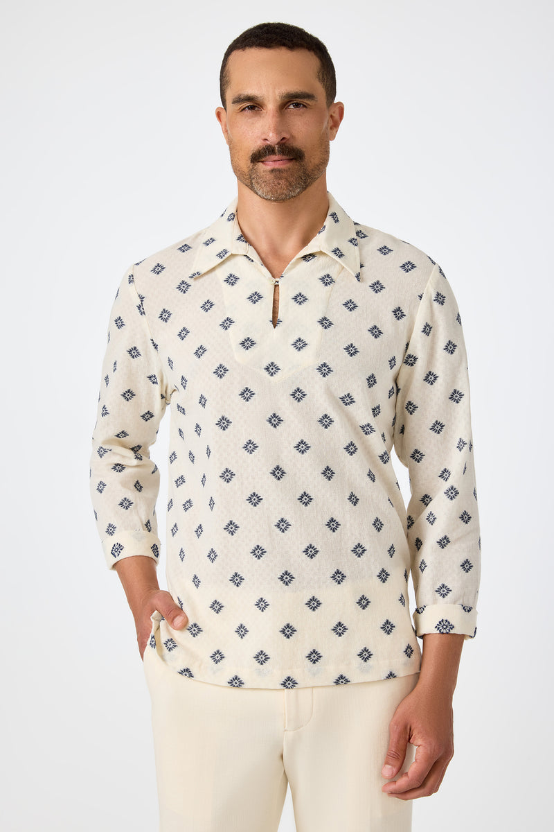 A man with short dark hair and a mustache wears the Hendrix Long-Sleeve Pullover in ecru, featuring a blue geometric pattern and a short zip at the collar, paired with light trousers, standing against a plain white background.