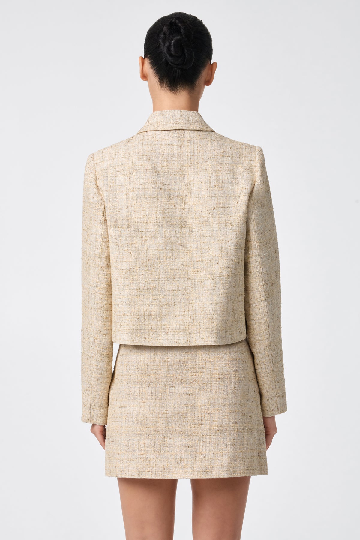 A woman stands with her back to the camera, wearing the Bayonne Jacket in beige with a matching skirt. Her hair is in a neat bun against a plain white background.