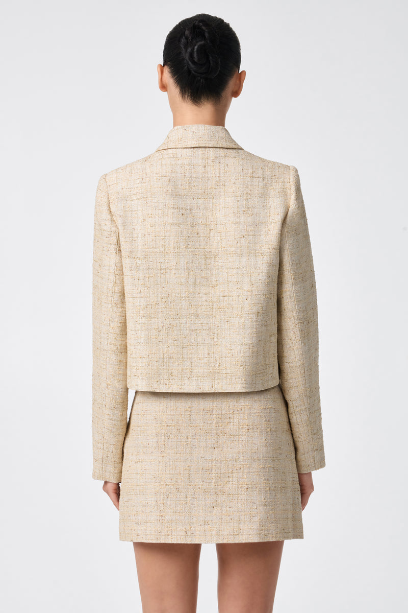 A woman stands with her back to the camera, wearing the Bayonne Jacket in beige with a matching skirt. Her hair is in a neat bun against a plain white background.