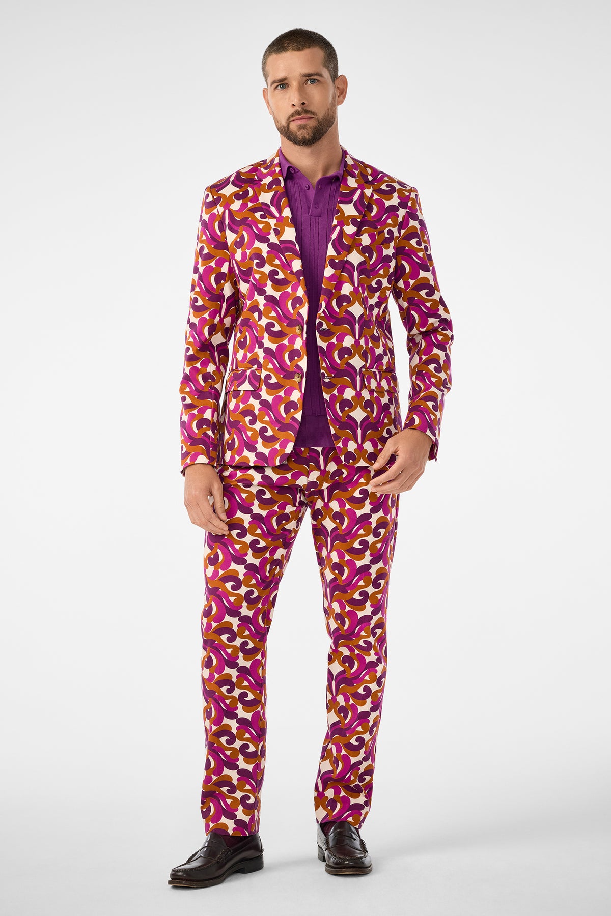 A man wears the Thurston Blazer, featuring a bold, colorful pattern in purple, magenta, yellow, and white, paired with a purple shirt and dark dress shoes against a plain white background.