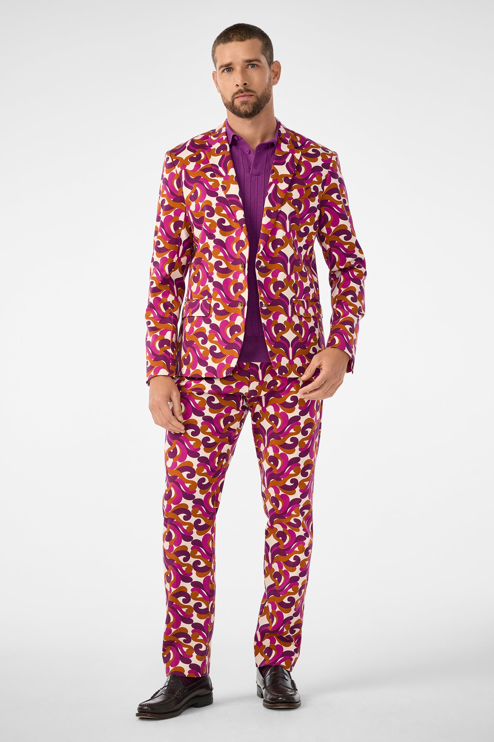 A man wears the Thurston Blazer, featuring a bold, colorful pattern in purple, magenta, yellow, and white, paired with a purple shirt and dark dress shoes against a plain white background.