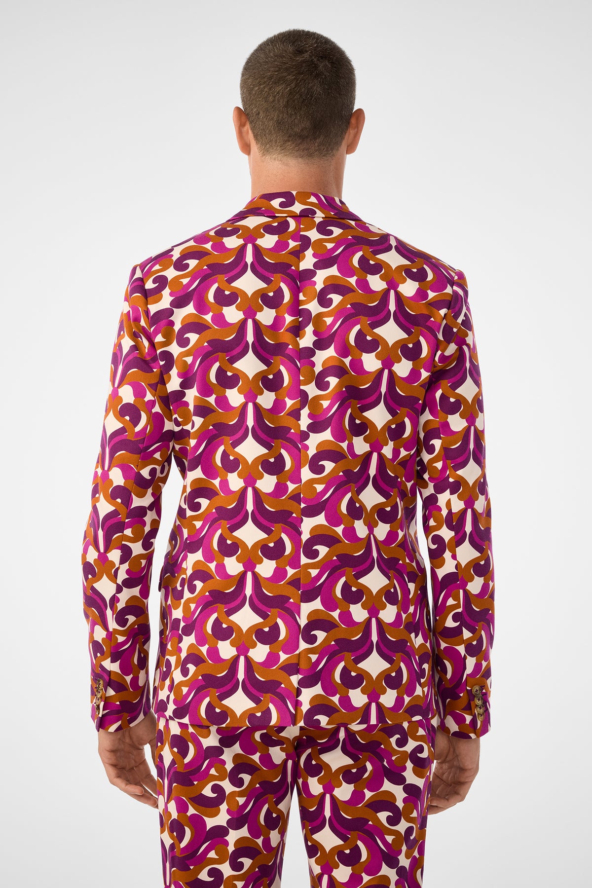 A man stands facing away, wearing the Thurston Blazer with matching trousers. Both feature a bold retro pattern in pink, purple, orange, and white with an intricate geometric design.