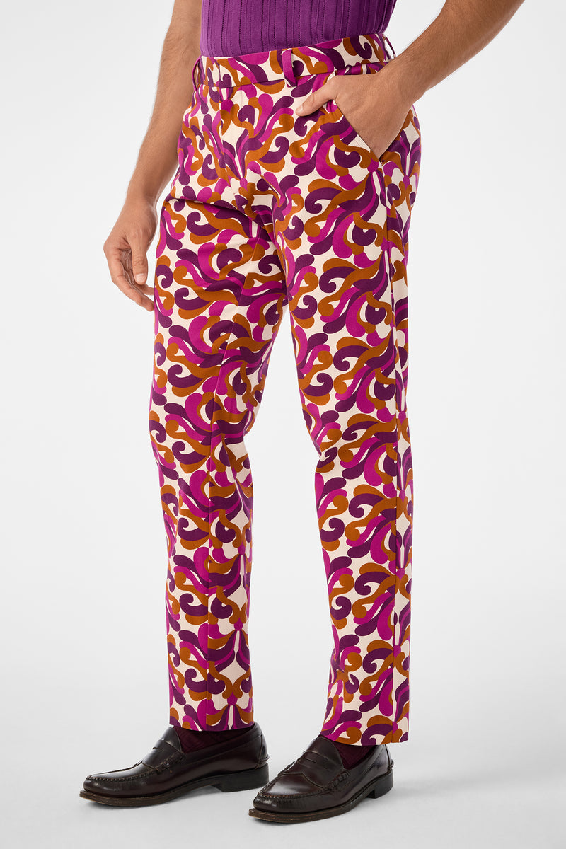 A person wearing Clyde Slim Trouser with swirling pink, purple, and yellow patterns, paired with a purple shirt and dark loafers, stands against a plain background with one hand in their pocket.