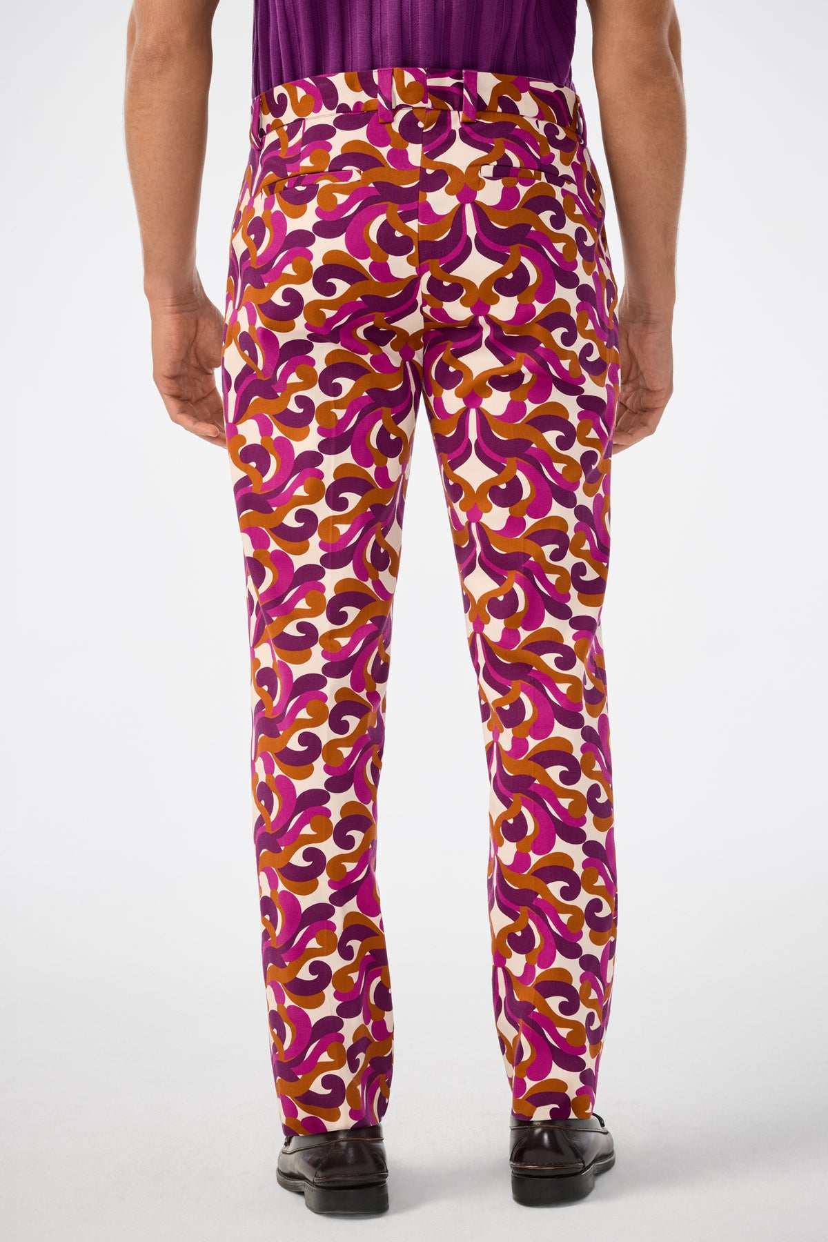 A person stands facing away, wearing the Clyde Slim Trouser featuring vibrant pink, orange, and white swirls, paired with a tucked-in purple shirt and black dress shoes against a plain white background.