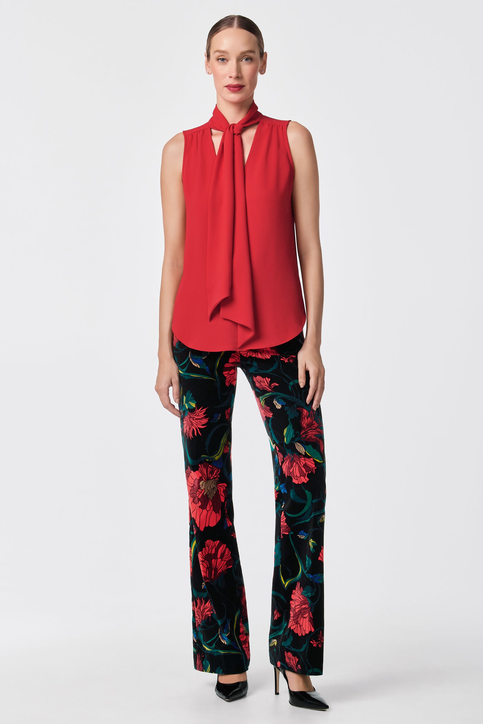 A woman faces forward in the Alora 2 Lavalliere Top, a sleeveless red blouse with a neck tie, paired with black pants in a bold red floral print and black high heels. The background is plain white.