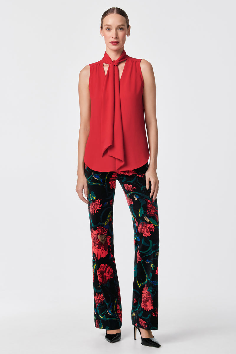 A woman faces forward in the Alora 2 Lavalliere Top, a sleeveless red blouse with a neck tie, paired with black pants in a bold red floral print and black high heels. The background is plain white.