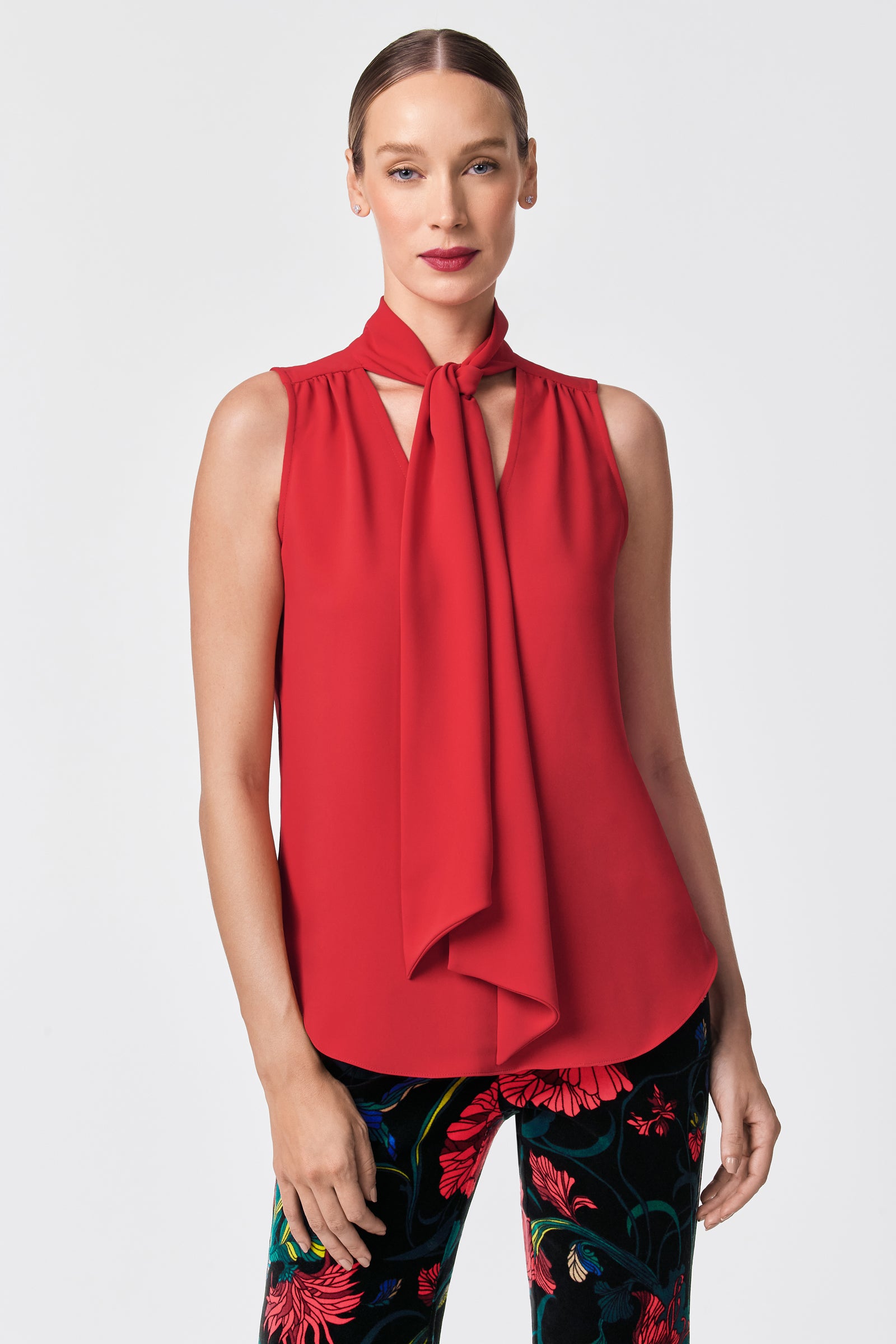 A woman wears the Alora 2 Lavalliere Top, a sleeveless red blouse with a necktie detail, paired with black pants featuring a bold floral pattern. Her hair is pulled back as she stands against a plain light background.