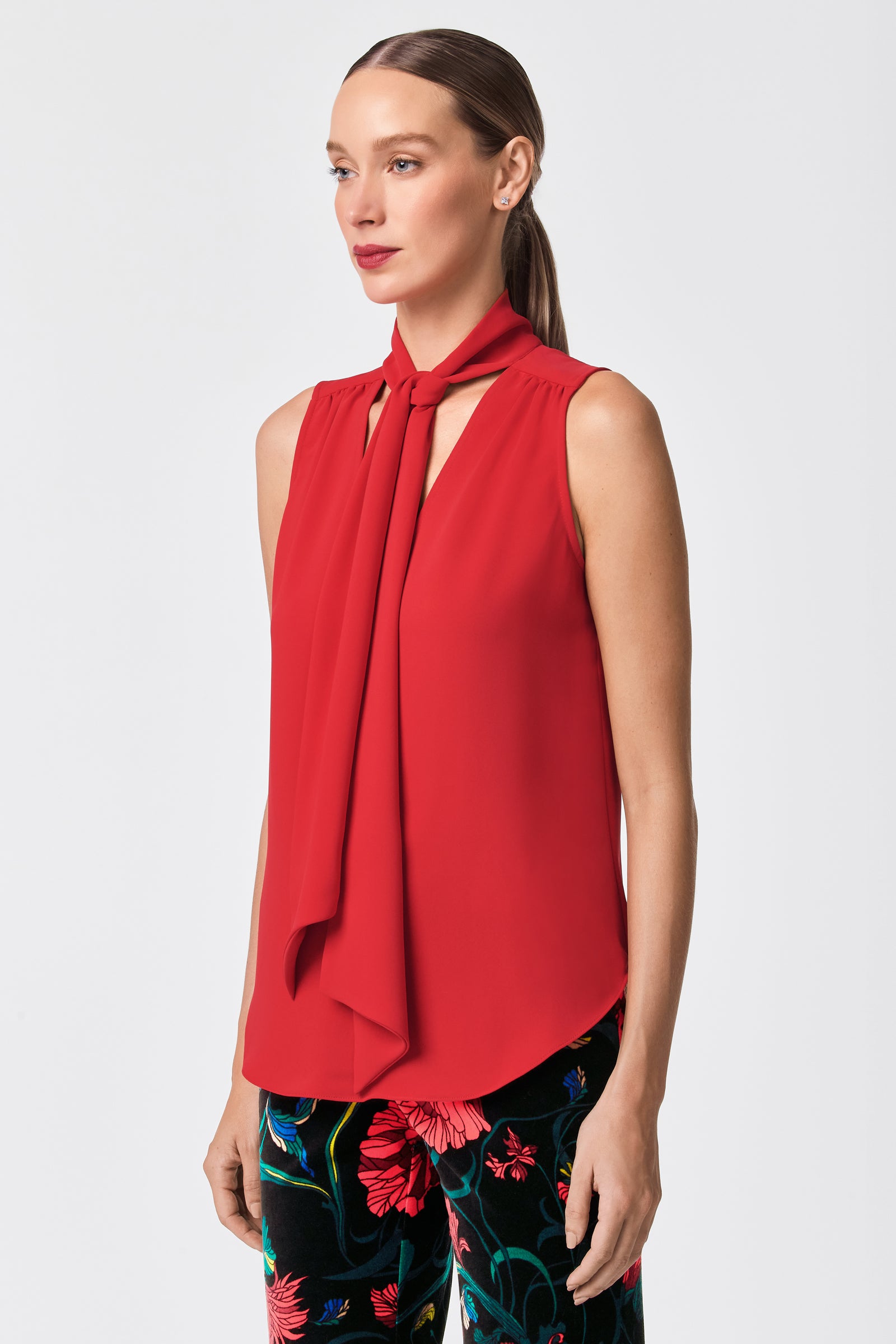 A woman wears the Alora 2 Lavalliere Top, a sleeveless red blouse with a front tie detail, paired with colorful floral pants. Her straight brown hair is pulled back as she gazes to the side against a plain light background.