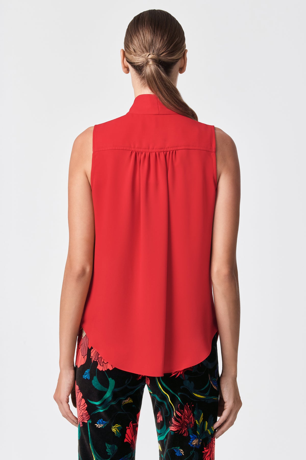 A woman is seen from behind wearing the Alora 2 Lavalliere Top, a sleeveless red blouse, paired with black floral pants. Her hair is styled in a low ponytail, and she stands against a plain white background.