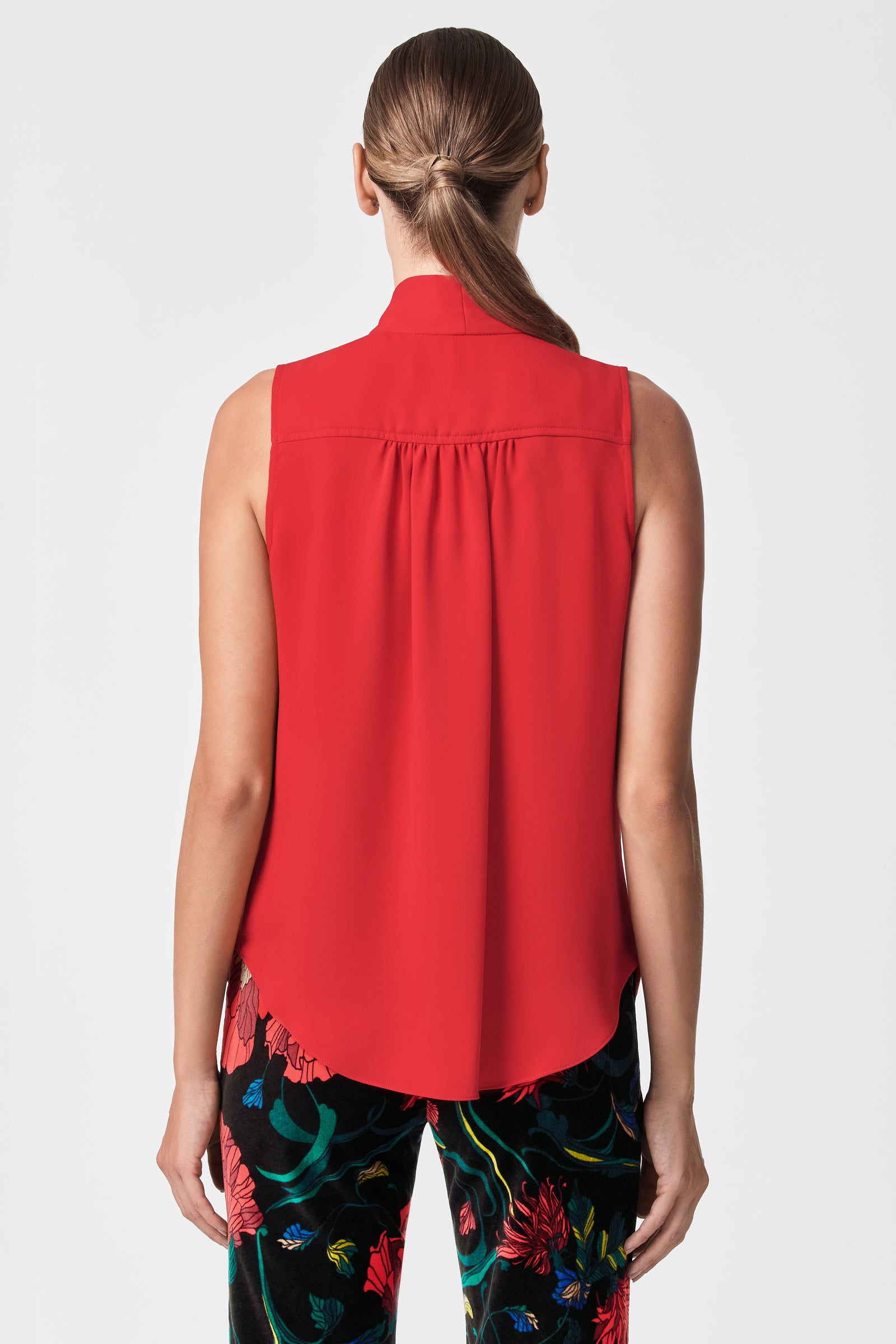 A woman is seen from behind wearing the Alora 2 Lavalliere Top, a sleeveless red blouse, paired with black floral pants. Her hair is styled in a low ponytail, and she stands against a plain white background.