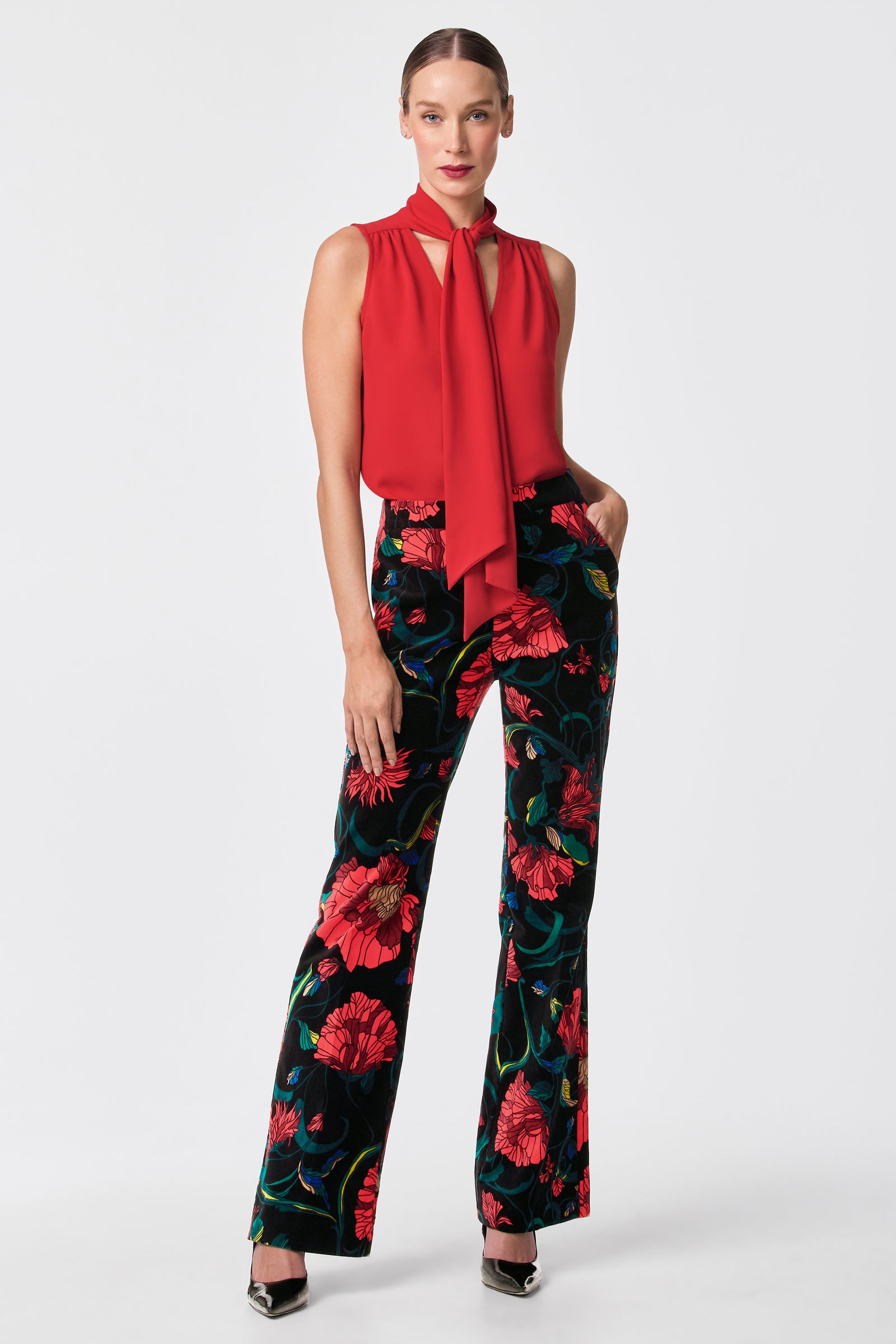 A woman stands against a white background wearing the sleeveless Alora 2 Lavalliere Top with a necktie and high-waisted black pants featuring a bold red floral pattern. Her hair is pulled back, and she finishes the look with silver pointed heels.