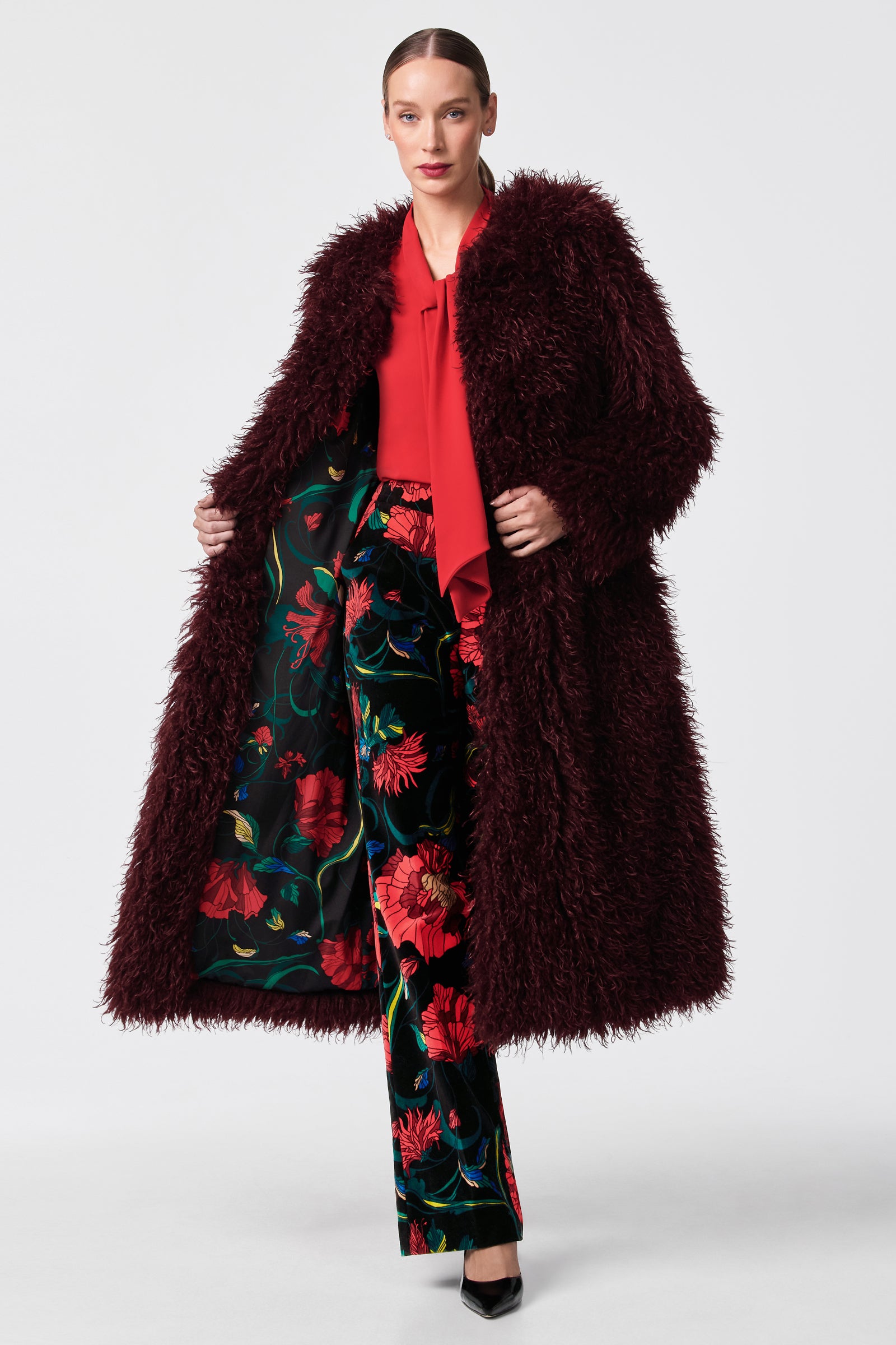 A woman models the Gladis Faux Fur Coat, a long burgundy piece, layered over a red blouse and dark green floral wide-leg pants with black heels, set against a simple light background.