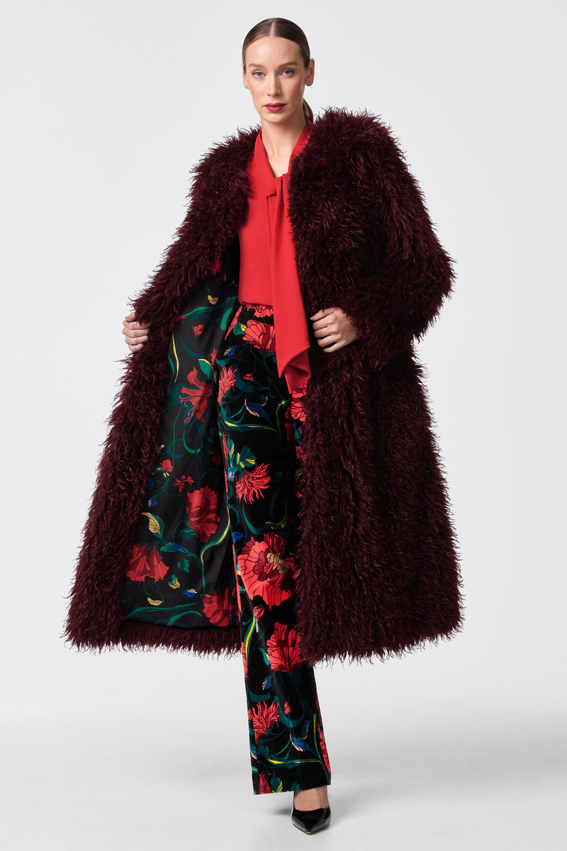 A woman models the Gladis Faux Fur Coat, a long burgundy piece, layered over a red blouse and dark green floral wide-leg pants with black heels, set against a simple light background.