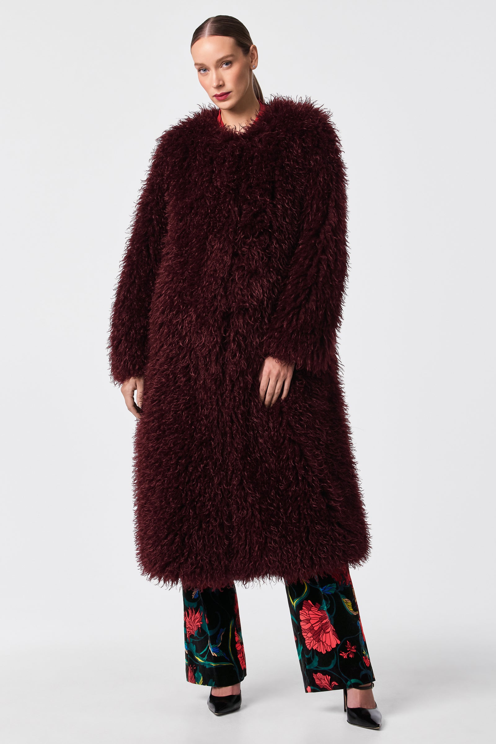 A woman stands against a plain white background, wearing the Gladis Faux Fur Coat in dark burgundy over black floral print pants and black pointed-toe heels.