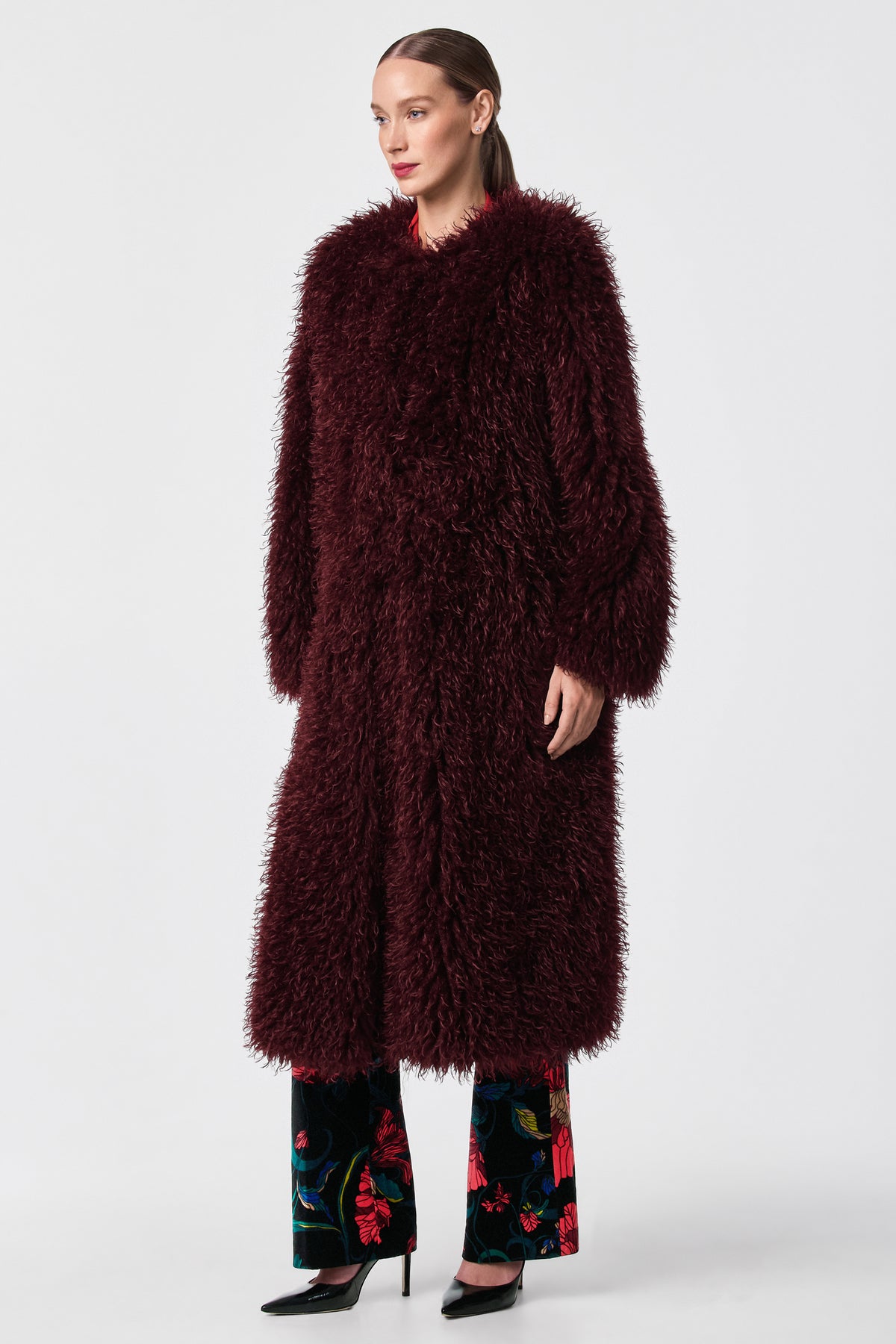 A woman stands against a plain background wearing the Gladis Faux Fur Coat in burgundy over floral pants and black heels. Her hair is tied back, and she gazes slightly to the side.