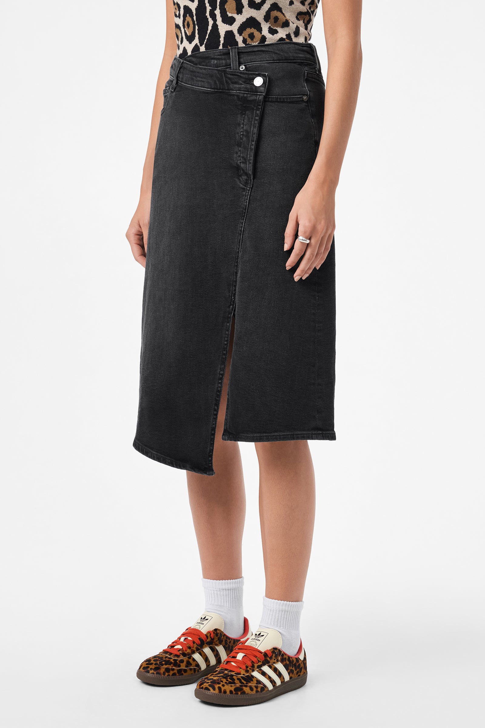 A person wearing a leopard print top, AG Bruna Skirt in black denim with a front slit, white socks, and animal print sneakers stands against a plain white background.