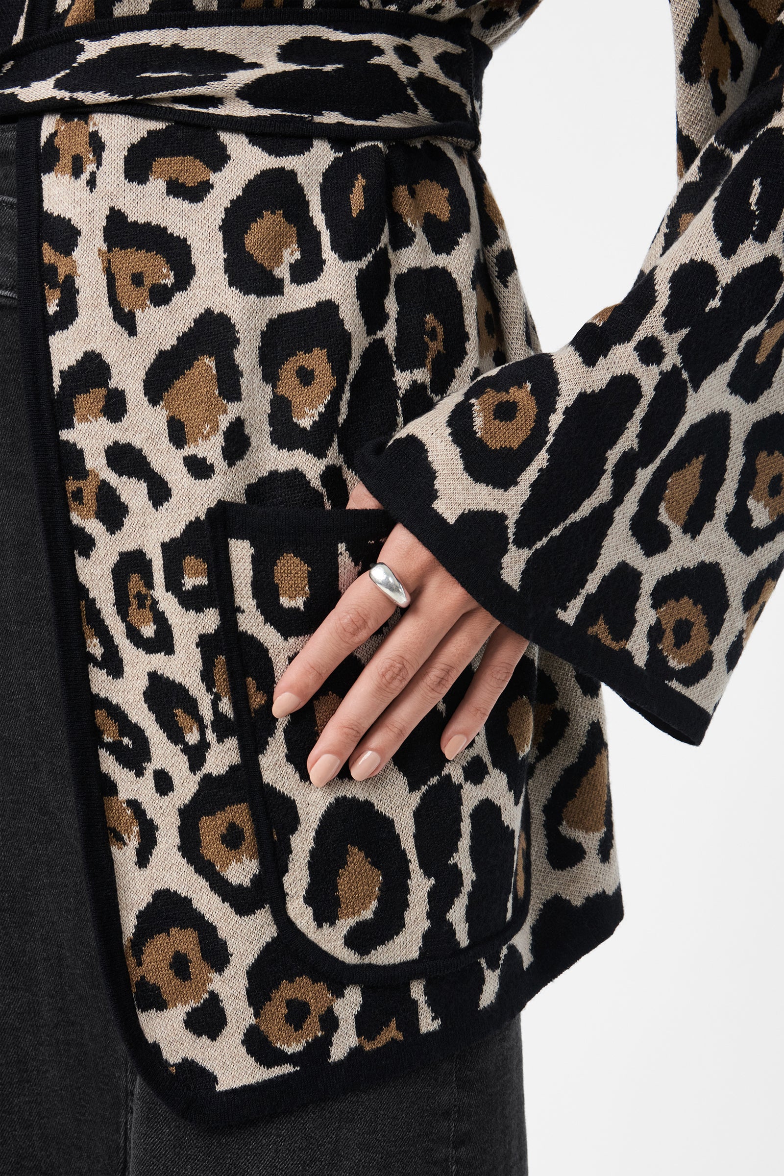 A close-up of a hand resting on the pocket of the Wright Cardigan, featuring a bold leopard print pattern. The person wears a silver ring and dark pants.