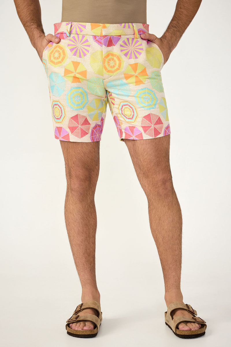 A person stands with hands in pockets against a plain background, wearing the Lawrence Short in Parapluie Jacquard, paired with a beige shirt and brown sandals.