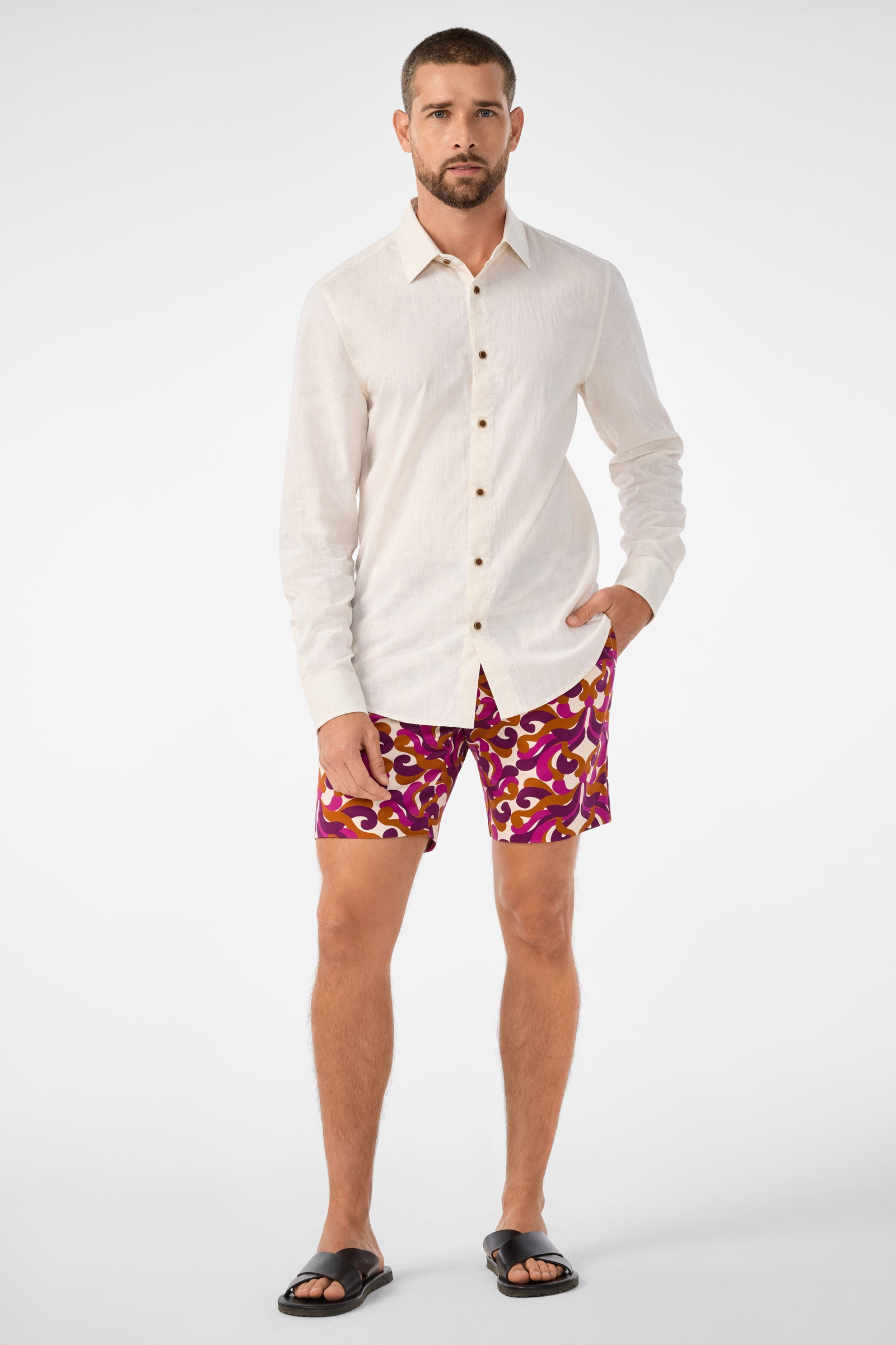 A man with short hair and a beard poses with one hand in his pocket, wearing the Lawrence Short—patterned magenta and yellow shorts—paired with a cream long-sleeve button-up shirt and black sandals against a plain background.