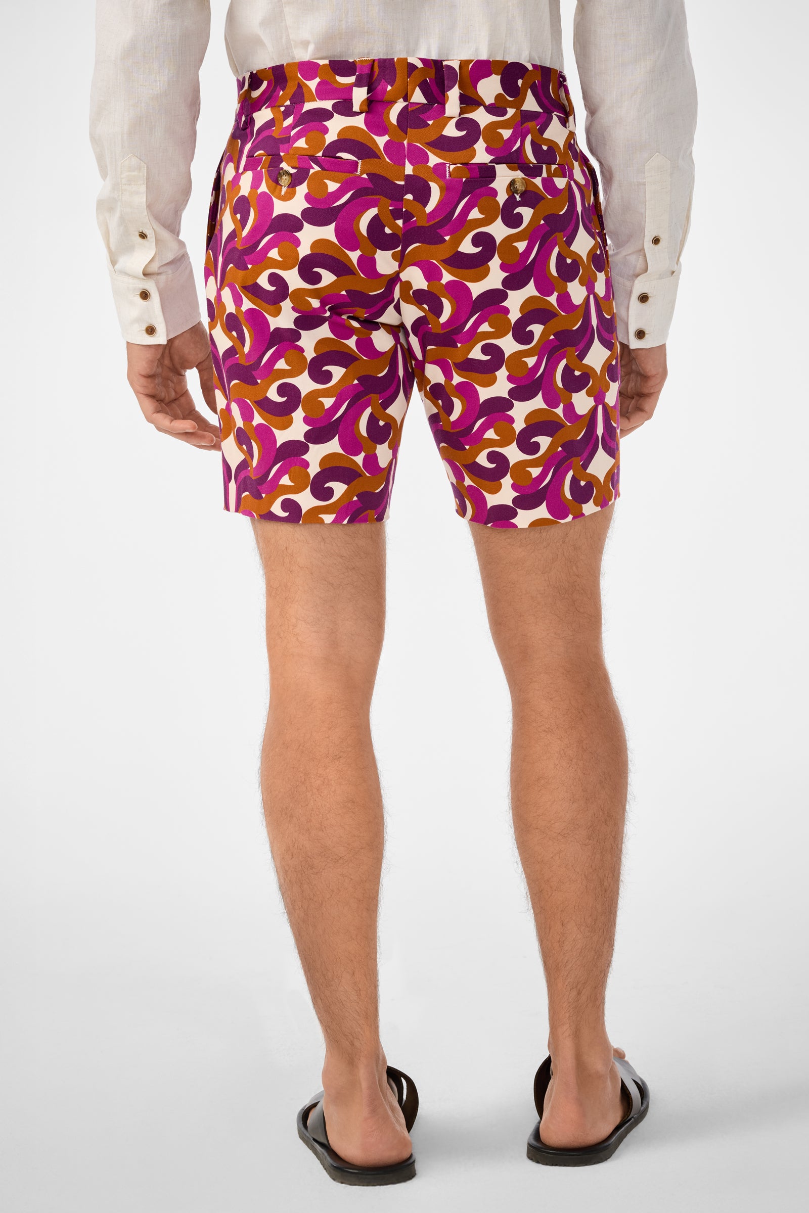 A person faces away, wearing a long-sleeve white shirt, black sandals, and vibrant Lawrence Shorts featuring pink, orange, and maroon patterns against a plain white background.