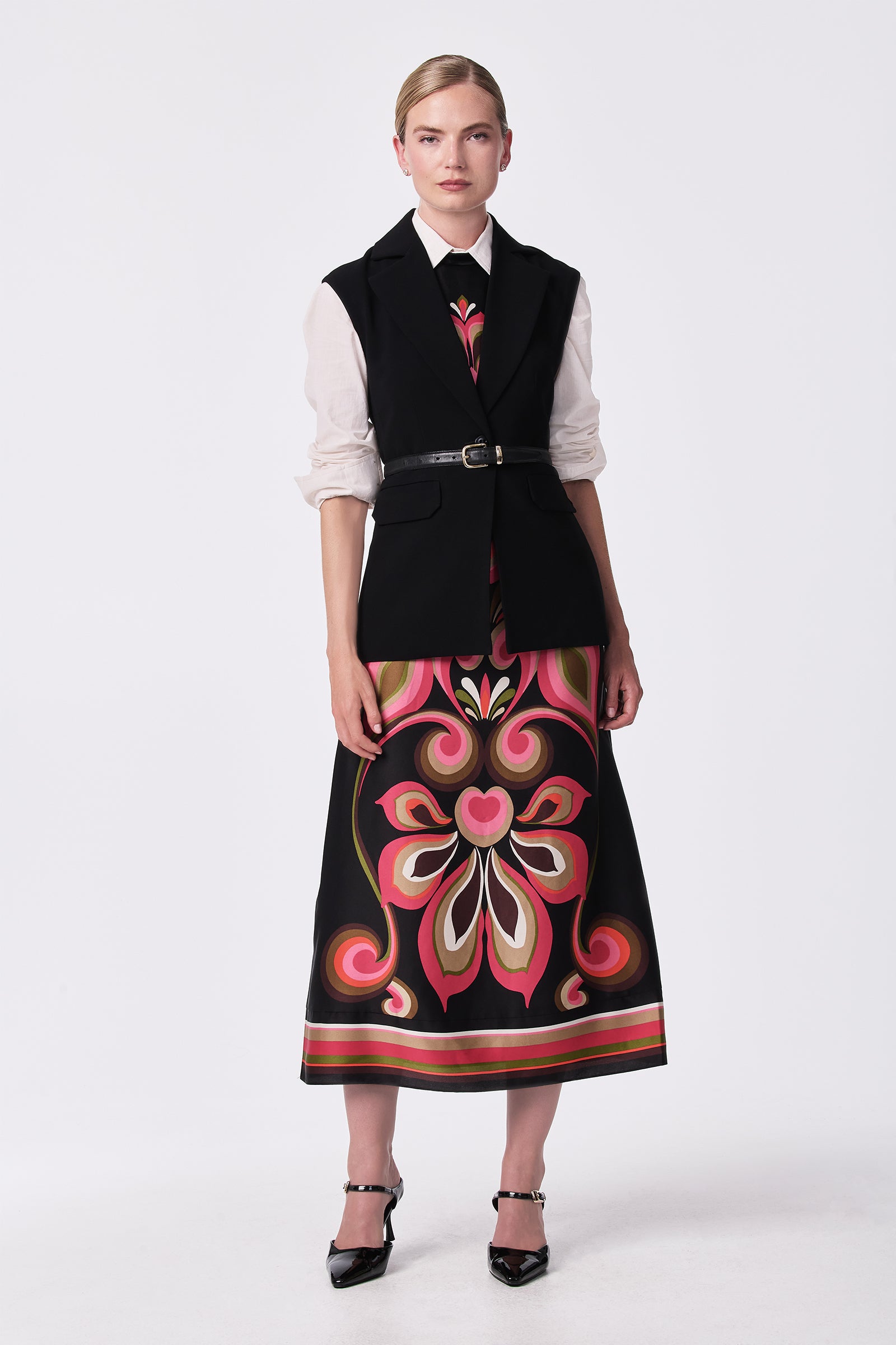 A woman stands against a plain white background wearing the Loveage Sleeveless Maxi Dress, featuring a black belted vest design and a long skirt with bold pink, red, and beige floral patterns. She has slicked-back hair and black heels.