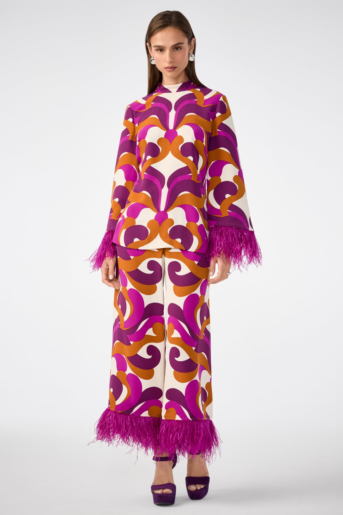 A woman wears the Short Weekend Pant featuring bold purple, orange, and cream swirling patterns with magenta feathered trim at the hem, paired with a matching top, purple platform heels, and statement earrings.