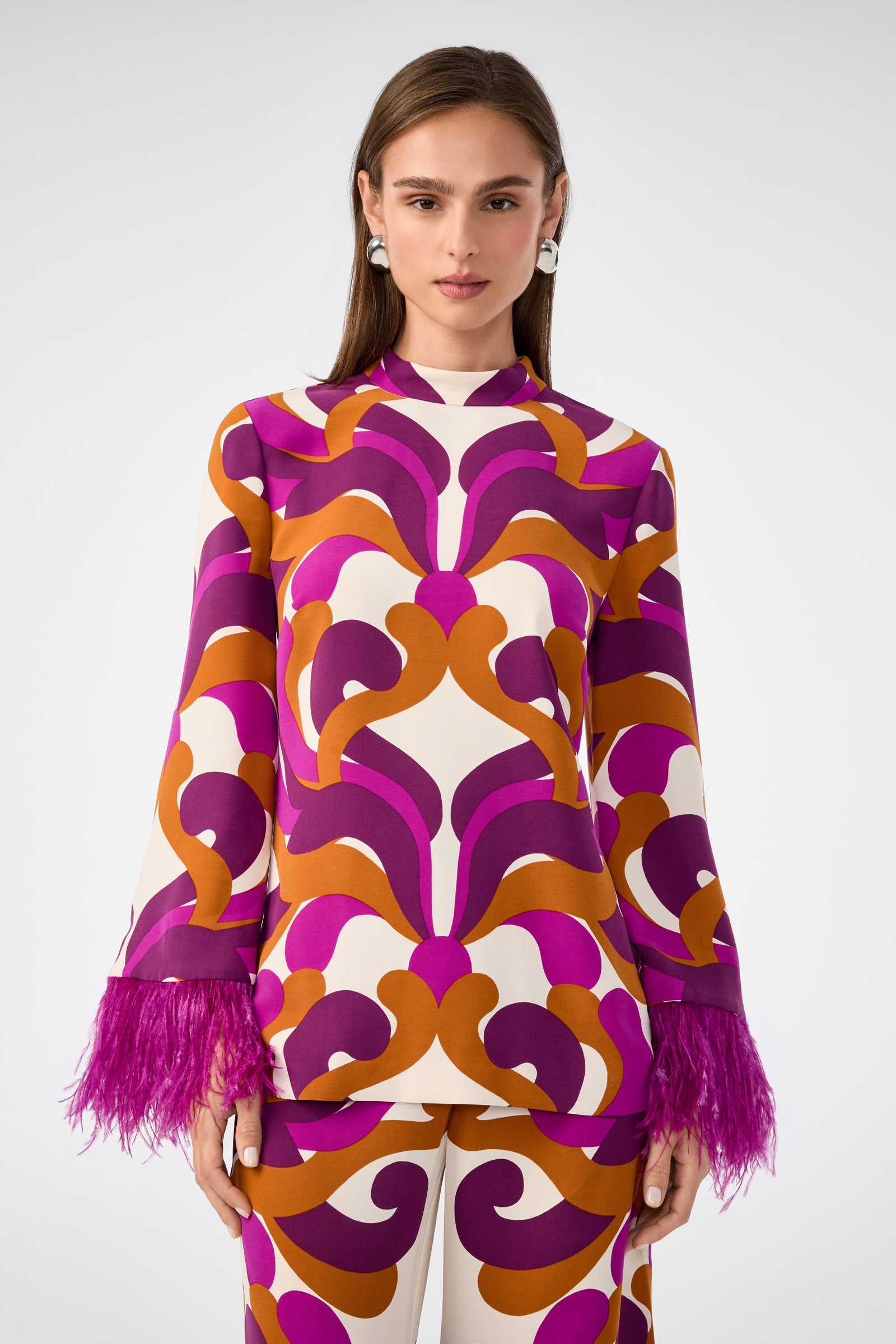 A woman with straight brown hair and earrings faces forward, wearing the Nyssa 2 Top—a colorful piece with bold purple, magenta, and orange geometric patterns and feathery magenta cuffs. She has a neutral expression.