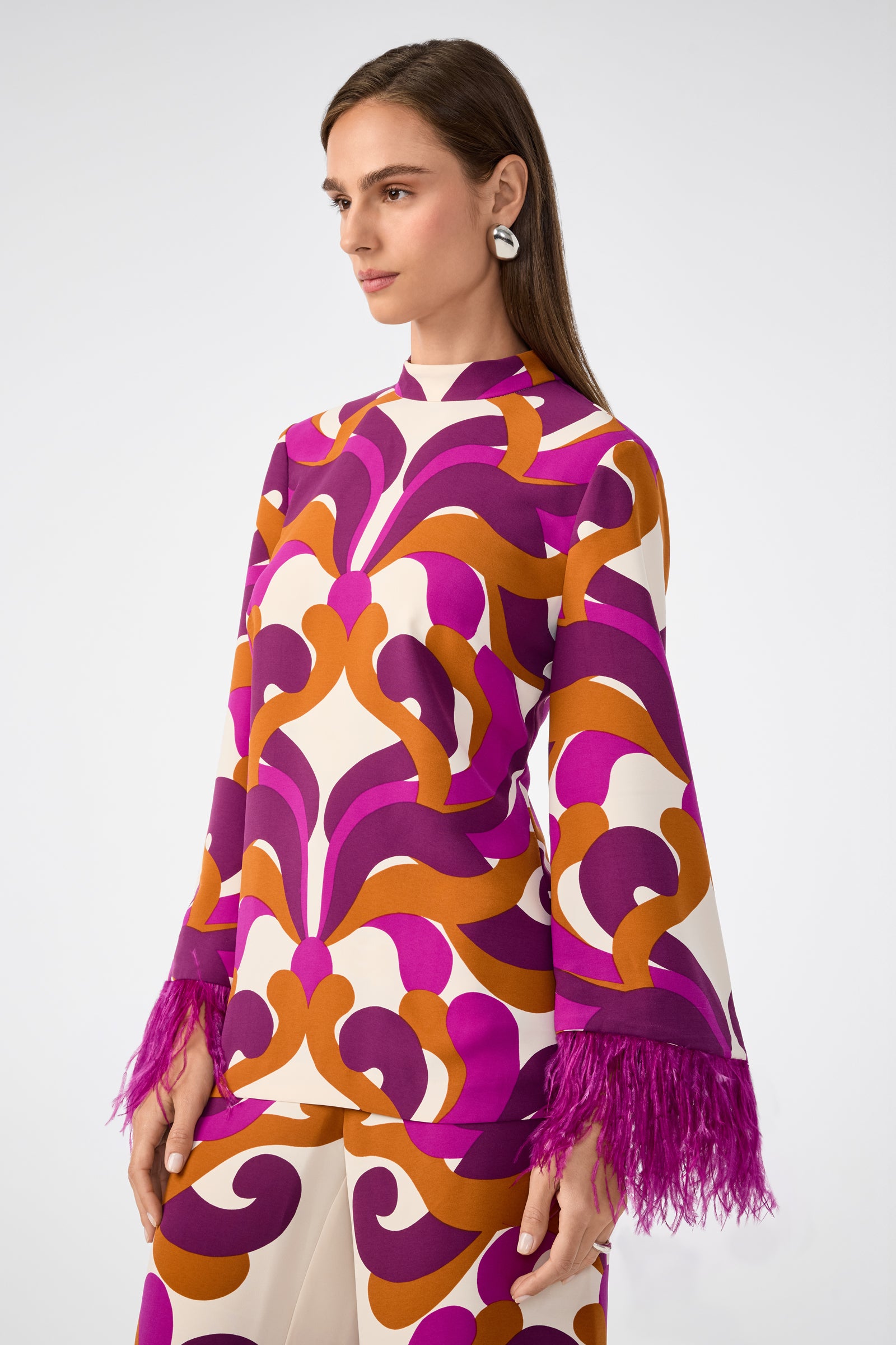 A woman with long brown hair wears the Nyssa 2 Top, featuring bold purple, orange, and white patterns and feathered sleeve cuffs. She accessorizes with silver statement earrings while standing against a plain background.