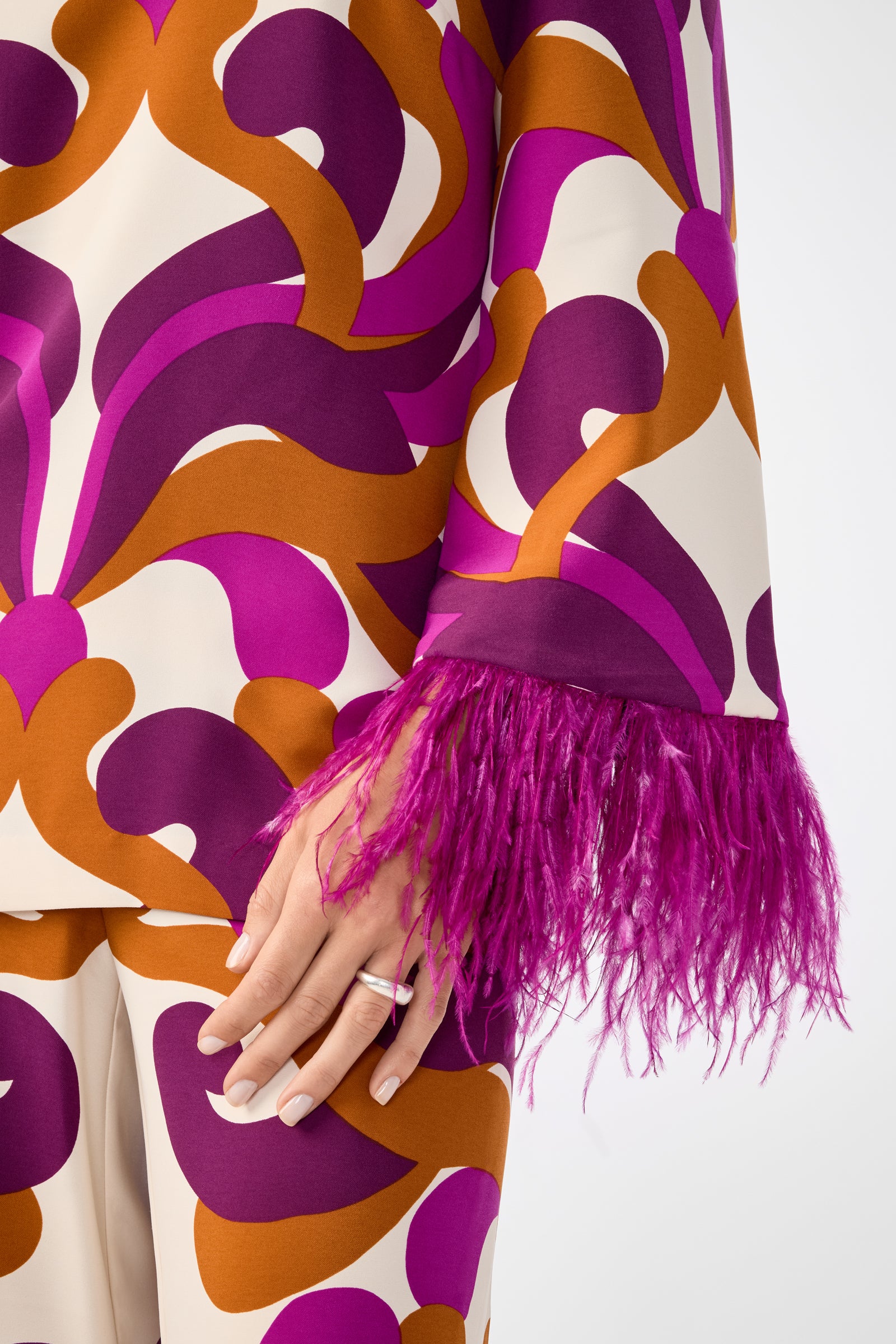 A person wears the Nyssa 2 Top, featuring bold purple, orange, and white patterns with bright magenta feather trim at the sleeve cuff. Their hand, adorned with a silver ring, is also visible.