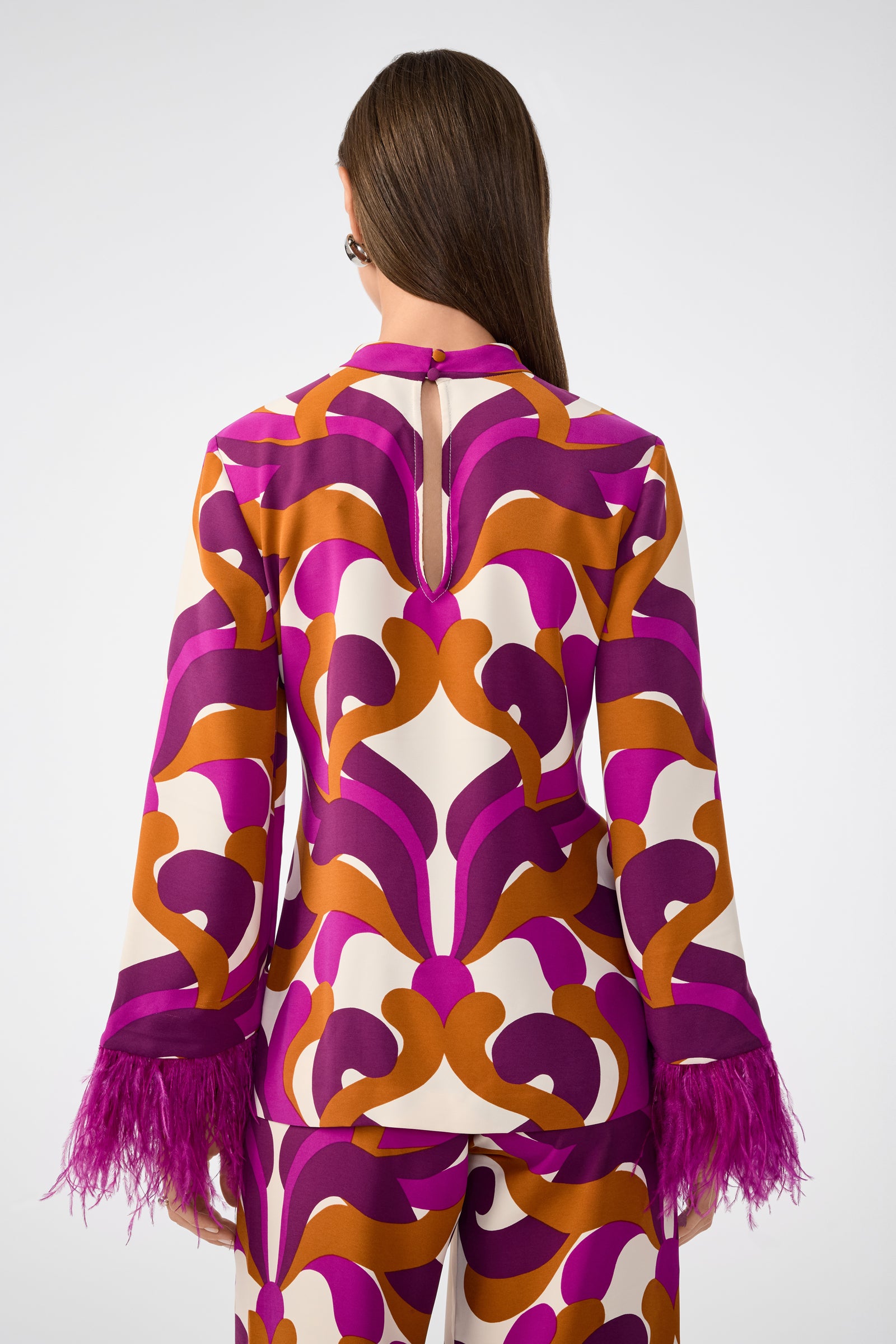 A woman is shown from the back wearing the Nyssa 2 Top, a long-sleeve blouse with bold abstract patterns in purple, orange, and white and finished with purple feathered cuffs.