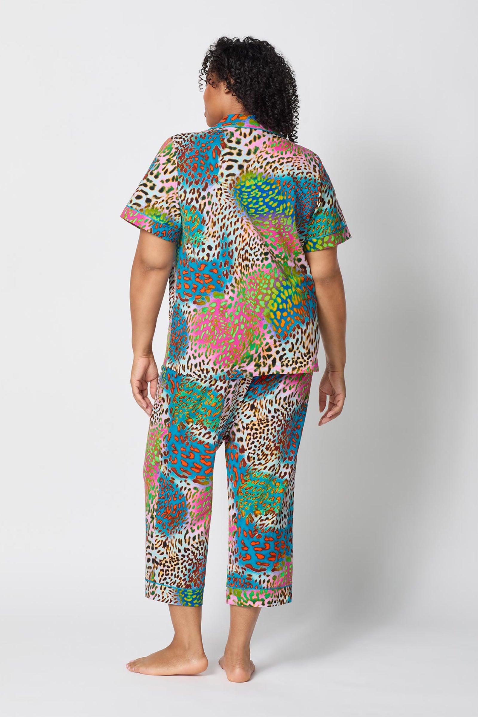 A person with curly hair stands barefoot facing away, wearing the Crop Classic PJ Set - Spray Leopard, a brightly colored short-sleeve pajama set with a bold pink, blue, green, and brown leopard print on white.