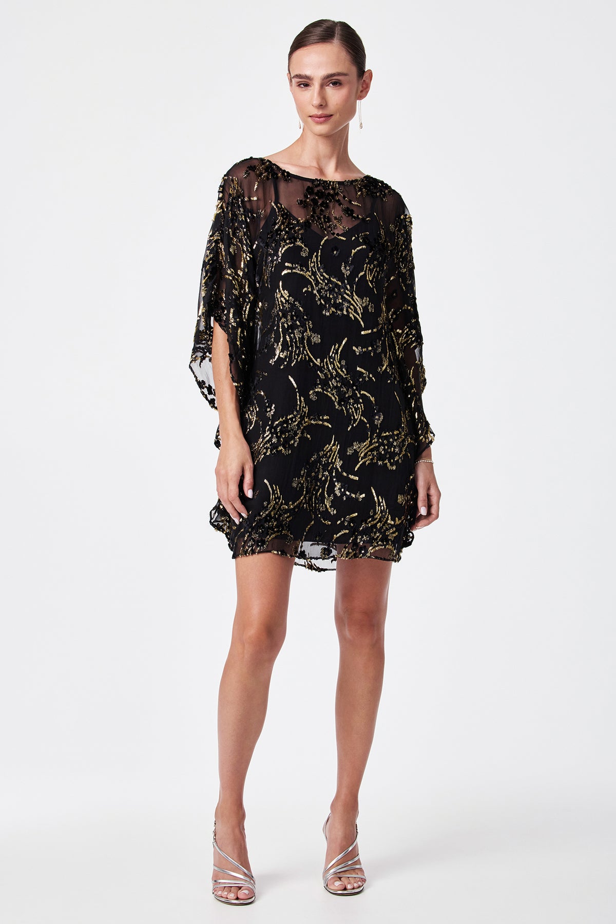 A woman poses against a plain white background wearing the Anissa Metallic Burnout Mini Caftan Dress with sheer black fabric and gold patterns, loose sleeves, silver strappy high heels, and her hair pulled back.