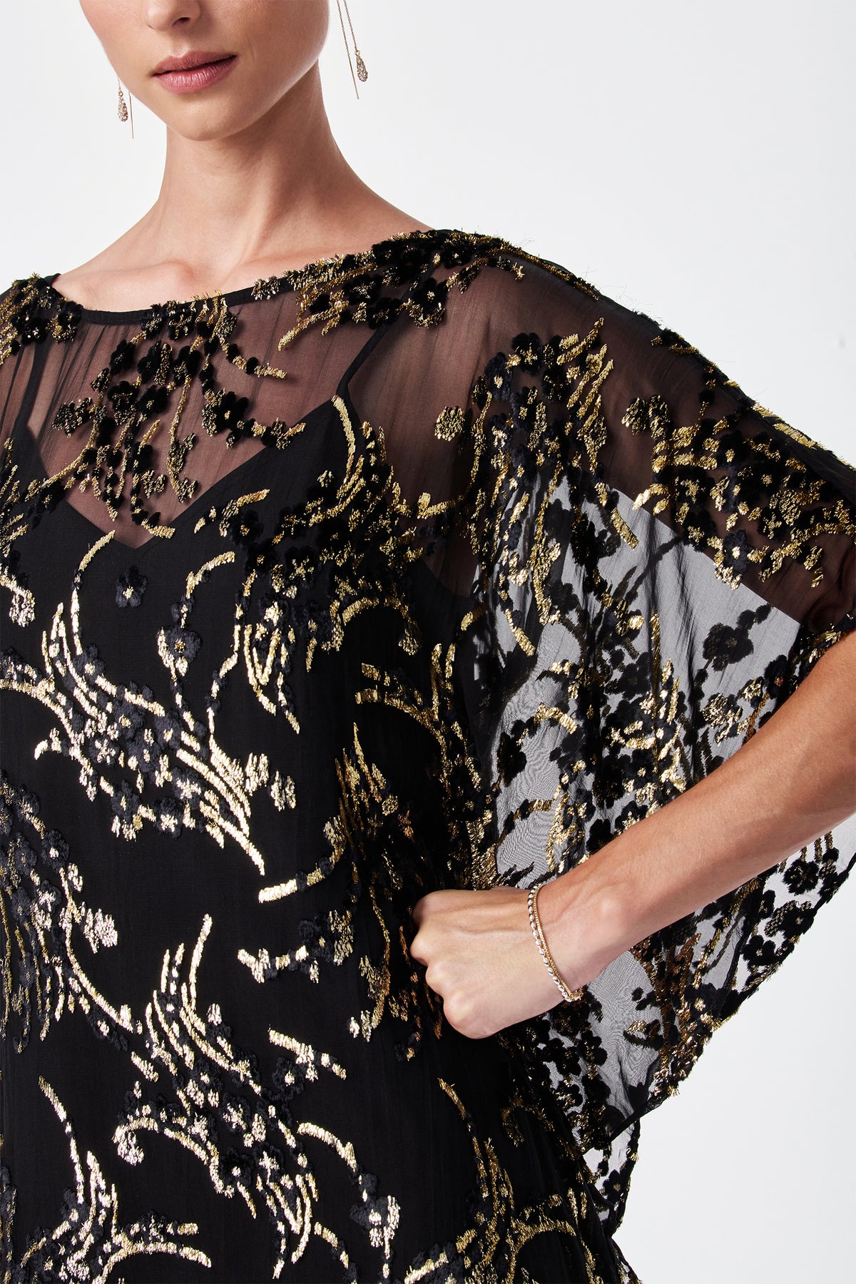 A woman models the Anissa Metallic Burnout Mini Caftan Dress, featuring sheer black fabric with intricate gold and black sequin embroidery. She poses with one hand on her hip; only her lower face and upper body are shown.