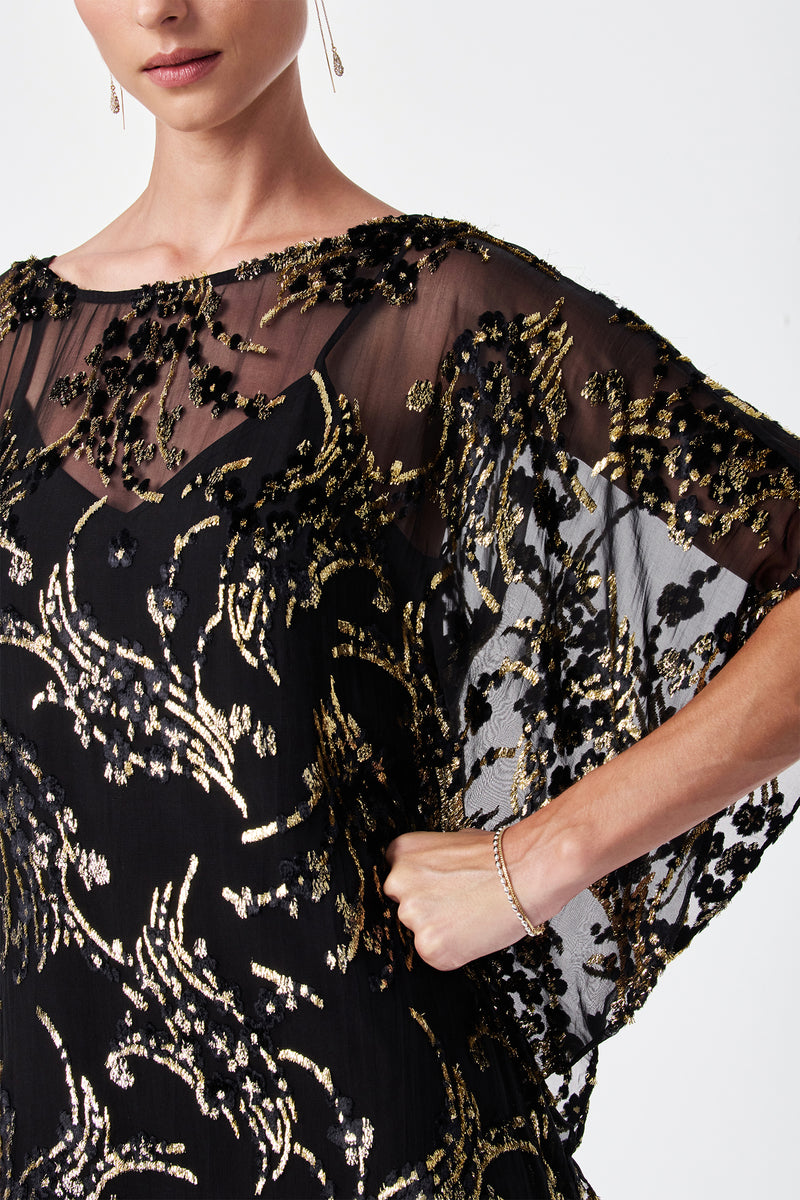 A woman models the Anissa Metallic Burnout Mini Caftan Dress, featuring sheer black fabric with intricate gold and black sequin embroidery. She poses with one hand on her hip; only her lower face and upper body are shown.