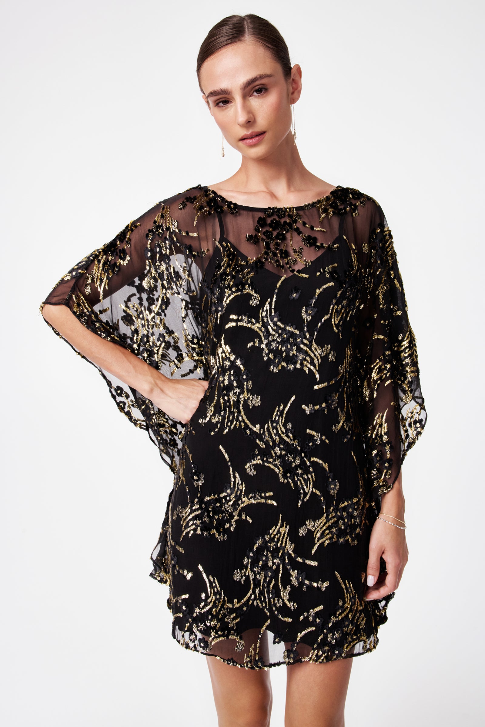 A woman stands with one hand on her hip against a white background, wearing the Anissa Metallic Burnout Mini Caftan Dress featuring sheer black fabric, gold sequin patterns, and loose, flowy sleeves.