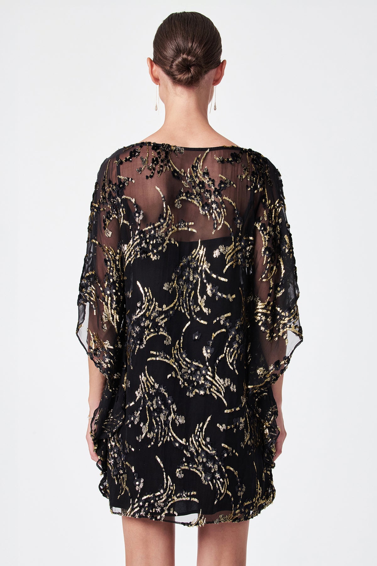 A person with dark hair in a neat bun is seen from the back wearing the Anissa Metallic Burnout Mini Caftan Dress, a short, loose black dress with gold and black sequin patterns, against a plain white background.