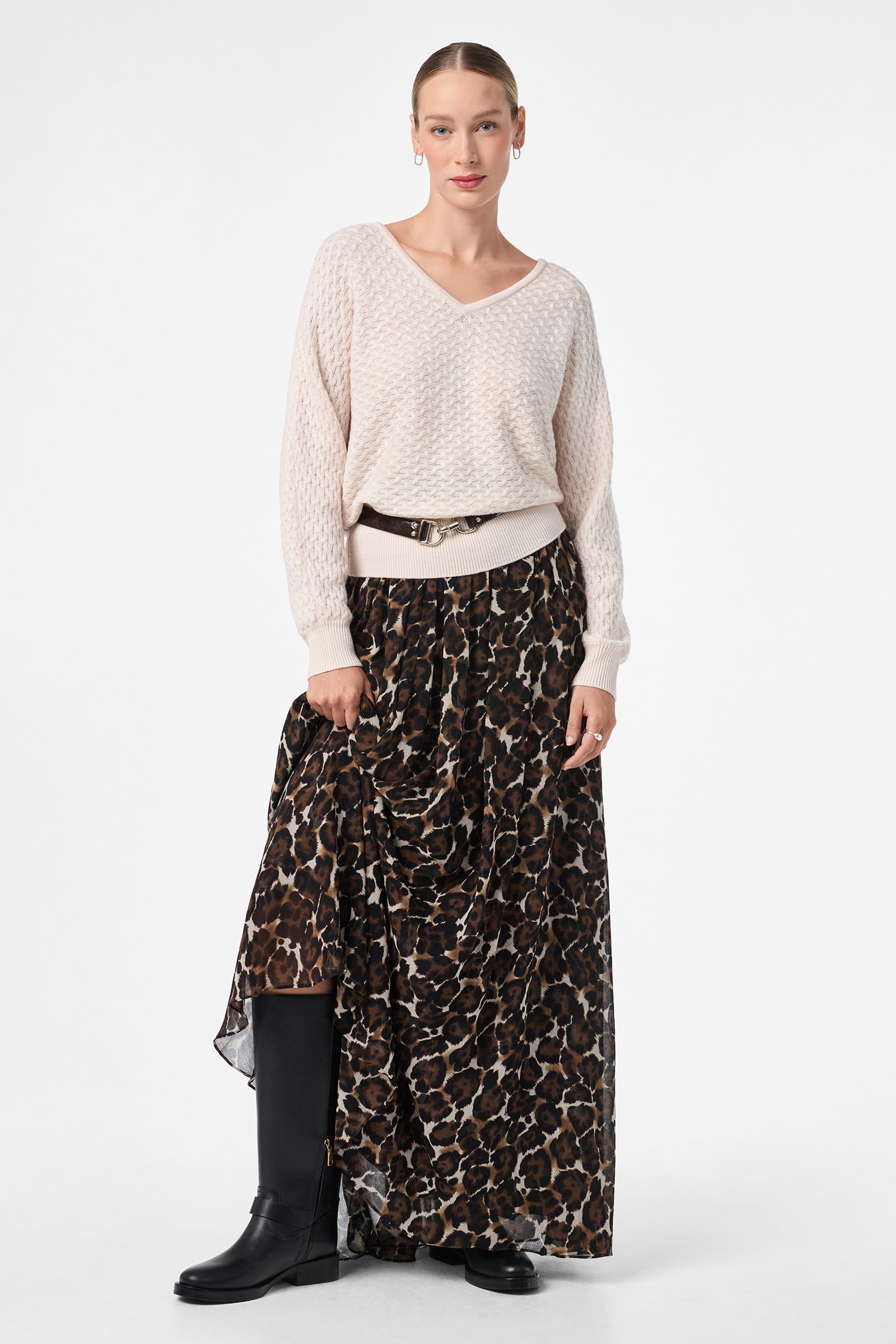A woman poses against a white background wearing the Bersha Skirt, a brown and black leopard print maxi, styled with a light knit sweater, black belt, and black knee-high boots. Her hair is pulled back as she holds her skirt with one hand.
