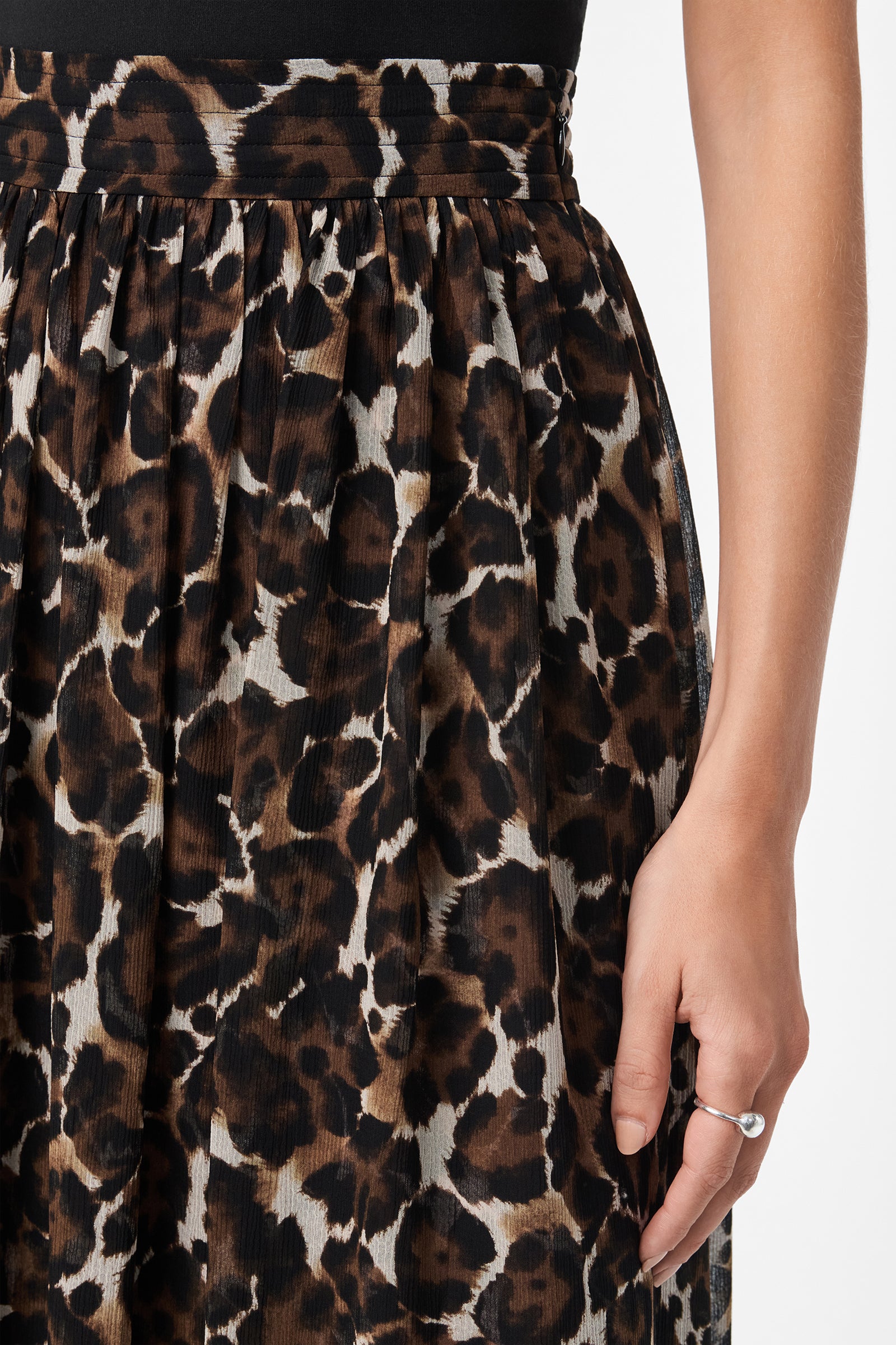 A person wears the Bersha Skirt, featuring brown, black, and white leopard print pleats, paired with a black top. A silver ring adorns their right hand as it rests by their side.