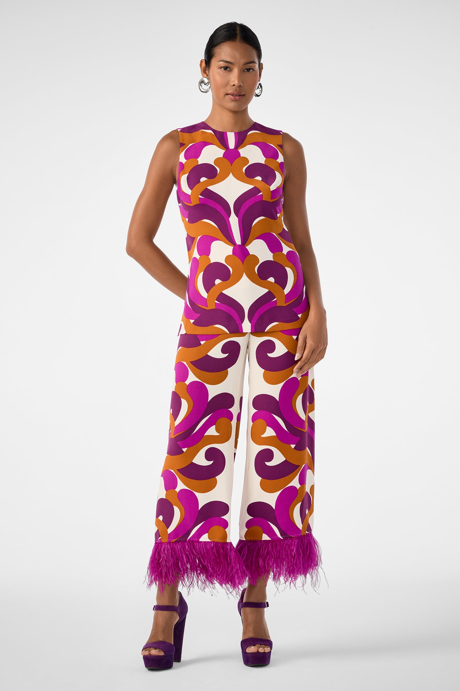 A woman models the Haruto Top—a sleeveless piece with a bold purple, pink, orange, and white 1970s-inspired pattern—paired with matching wide-leg pants with feather-trimmed hems and purple platform heels against a plain background.