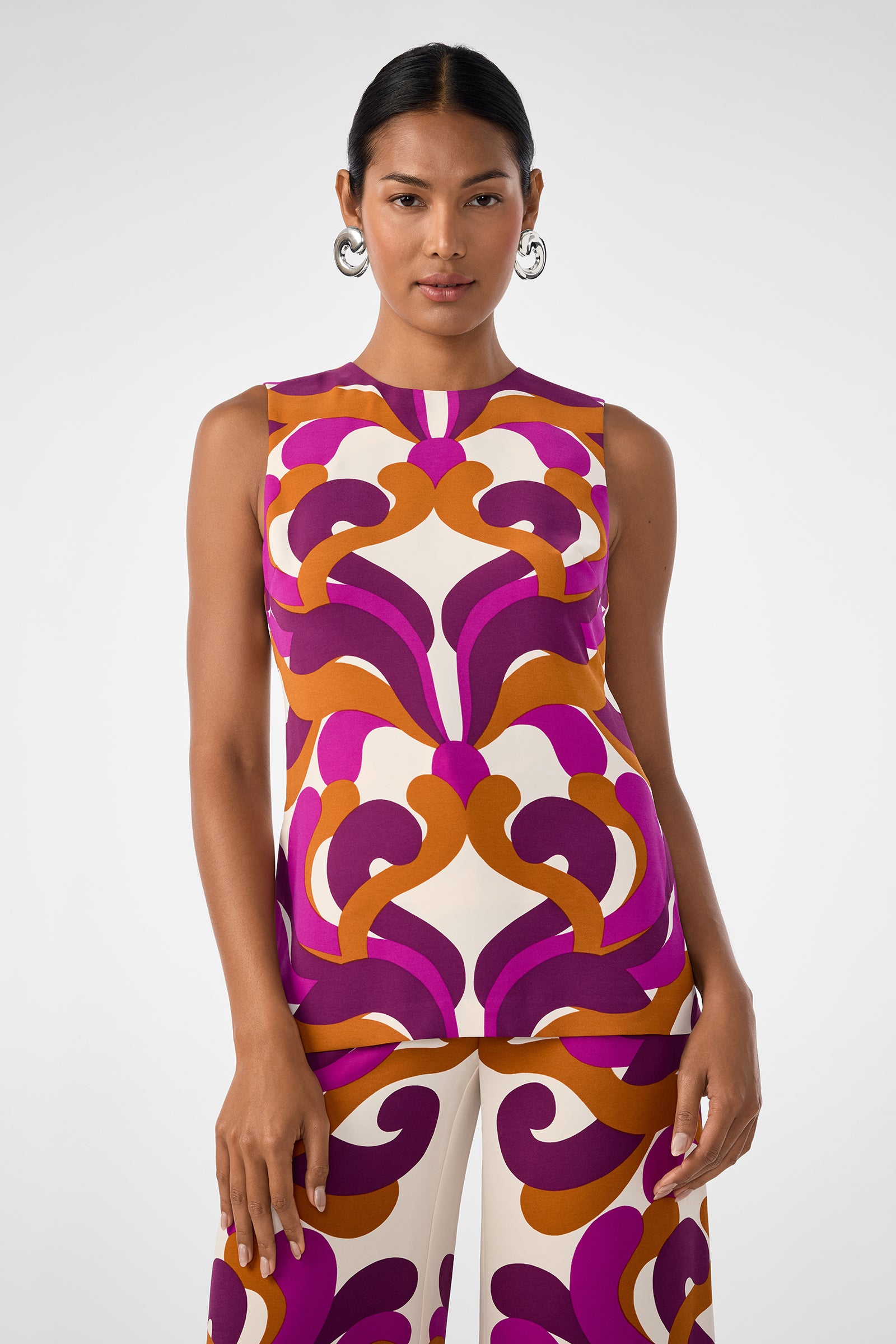 A woman wears the Haruto Top, a sleeveless top with a bold geometric pattern in purple, orange, and cream. She pairs it with matching pants, has her hair pulled back, and accessorizes with hoop earrings against a light background.