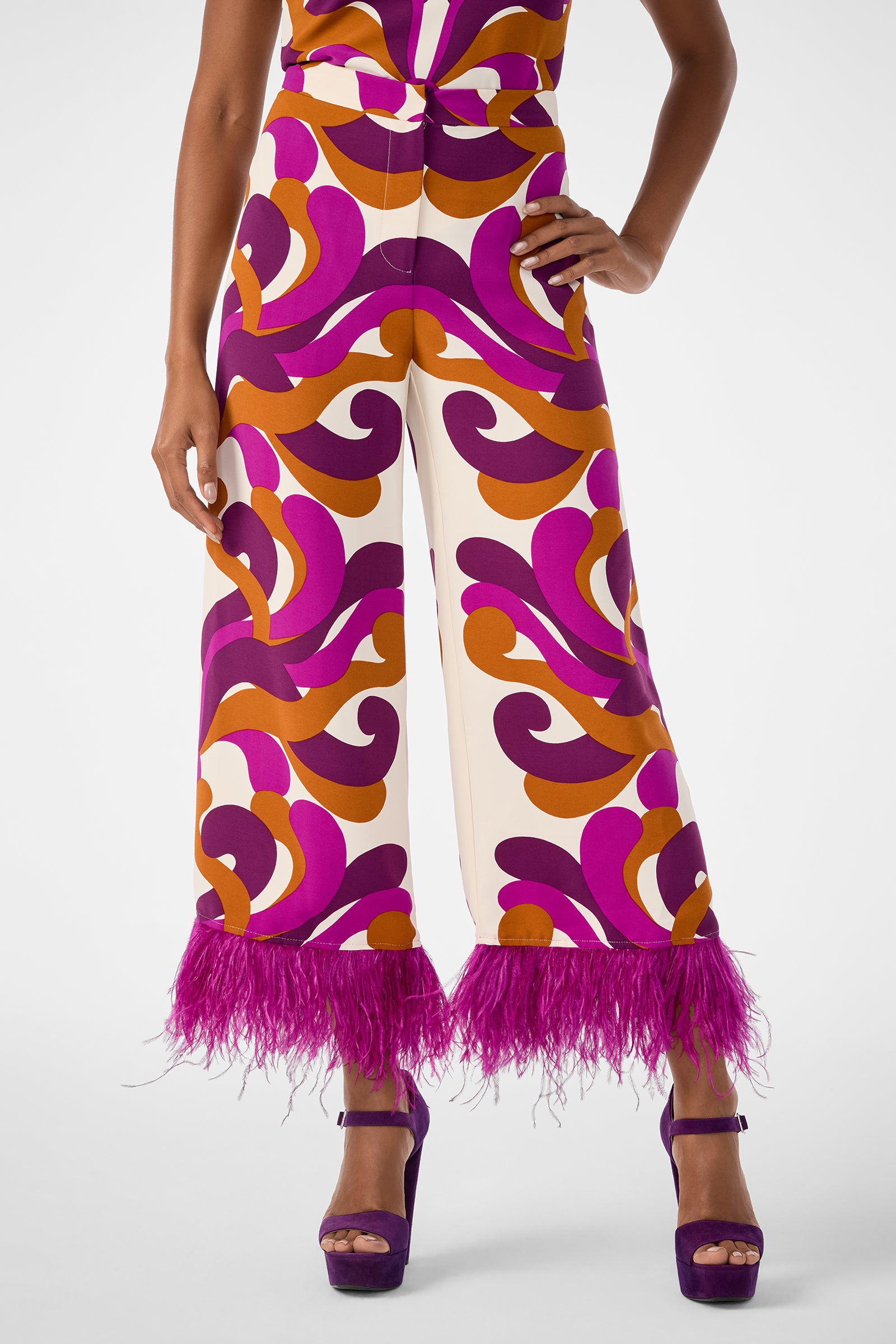 A person wearing the Short Weekend Pant, featuring a bold purple, orange, and white swirl print with pink feather trim at the hem, styled with purple platform heels and one hand on their hip.