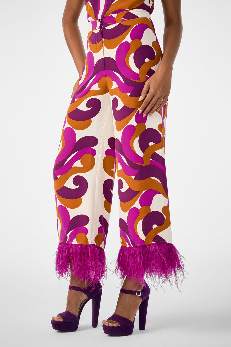 Wearing the Short Weekend Pant, featuring high-waisted, wide-leg style with a vibrant orange, purple, and pink swirl pattern and purple feather-trimmed hems, paired with purple platform heels.