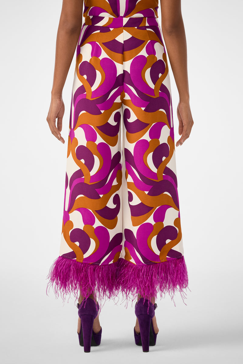 A person faces away, wearing the Short Weekend Pant—high-waisted, wide-leg with bold purple, orange, and cream swirls and purple feather hem—paired with purple high heels.