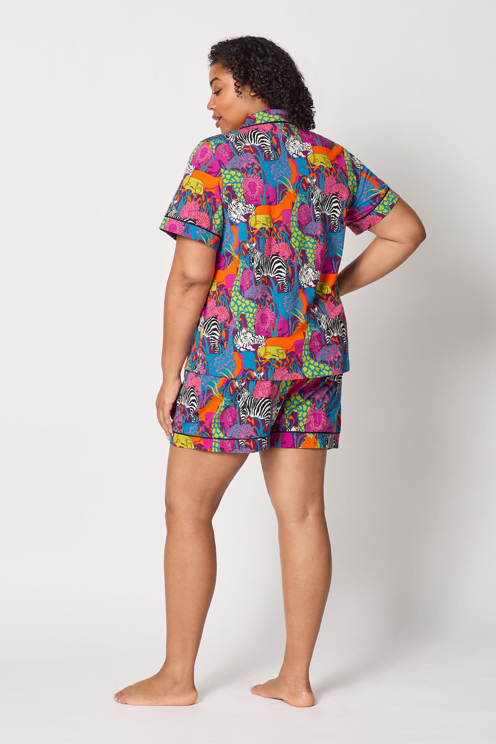 A person stands barefoot, facing away, wearing the Shorty PJ Set - Jungle: a colorful short-sleeve button-up pajama top and shorts with bright zebra and giraffe prints on a white background.